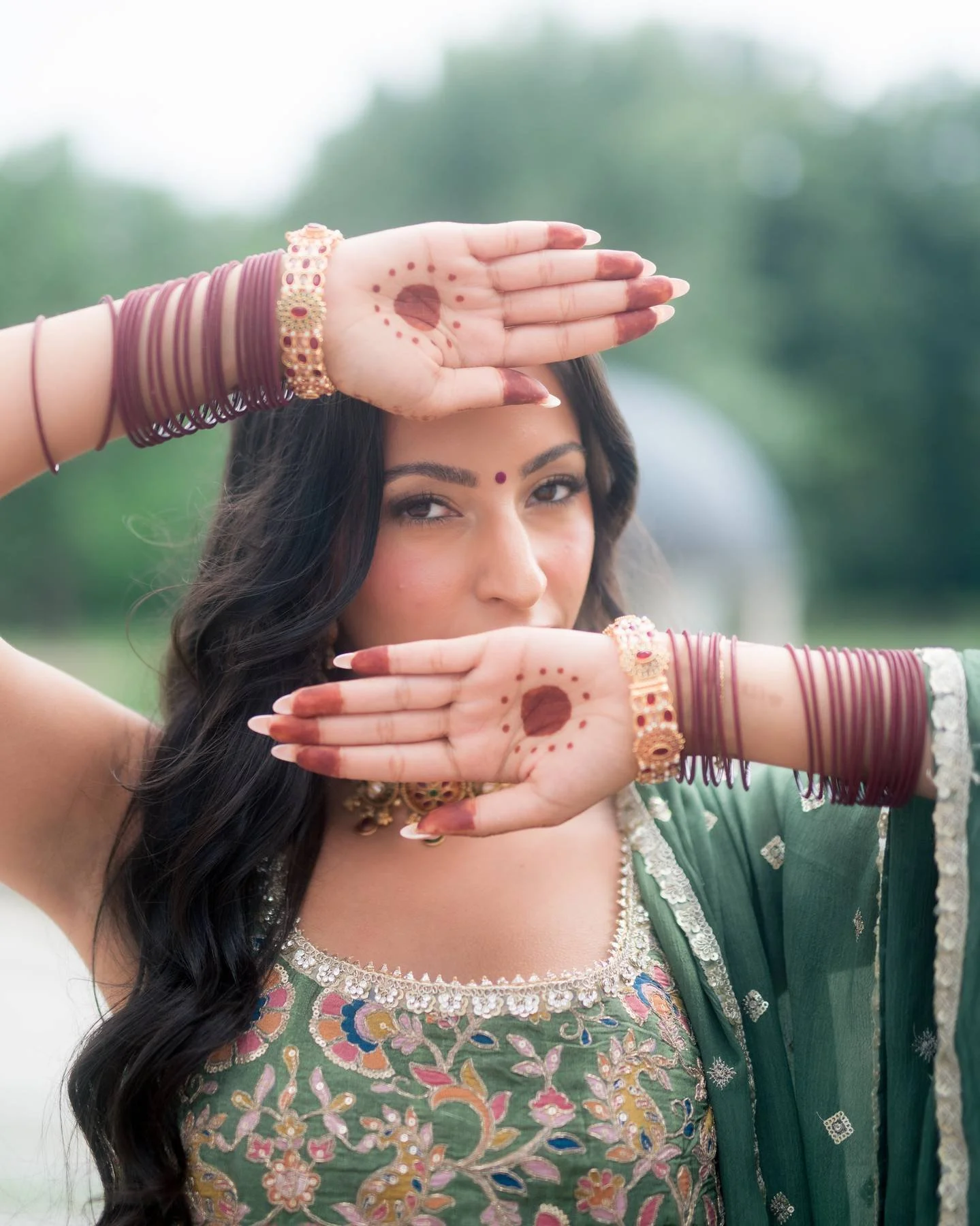 A woman dressed in traditional Indian attire with decorative jewelry, has henna designs on her hands, and is posing with her hands covering her forehead and mouth, outdoors with greenery in the background.