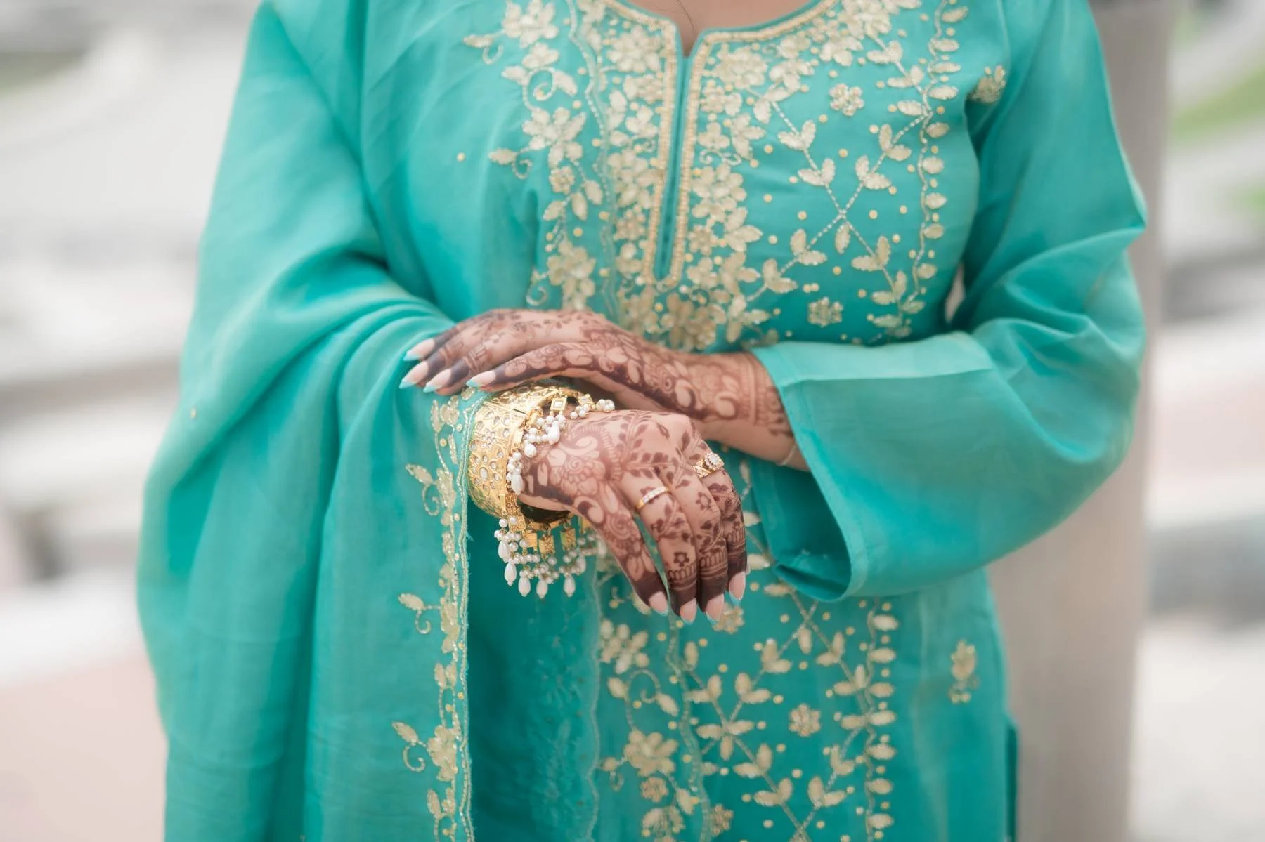 Close-up of a woman wearing a turquoise traditional dress with gold embroidery, displaying her hands with henna tattoos and gold jewelry.