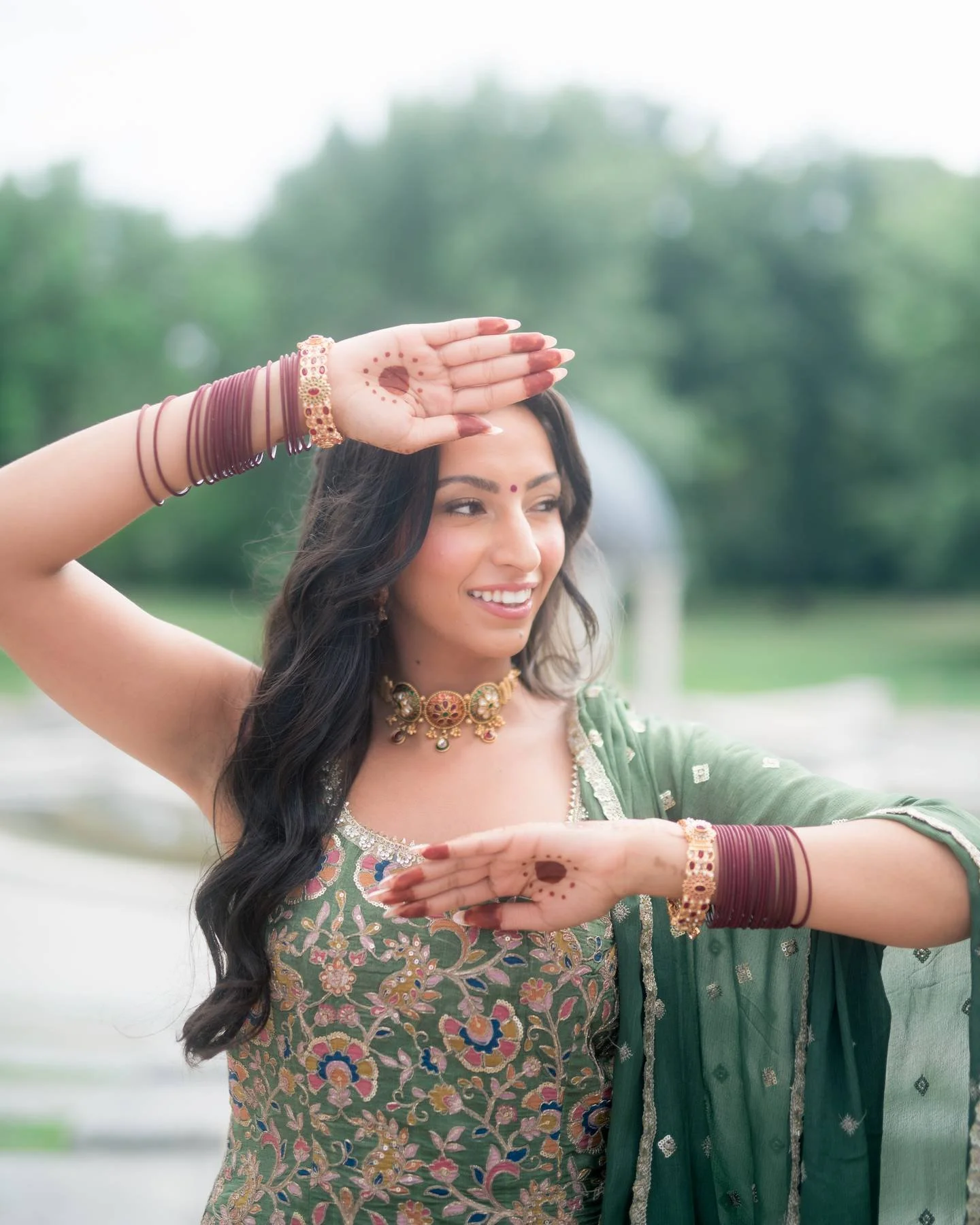 Woman dressed in traditional Indian attire performing a dance outdoors, with decorative jewelry and henna on her hands.