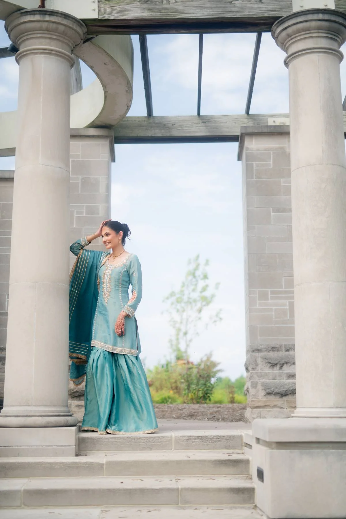 A woman dressed in a traditional turquoise Indian outfit, standing on stone steps between two large columns under a partially constructed stone archway, with a blurred outdoor landscape in the background.