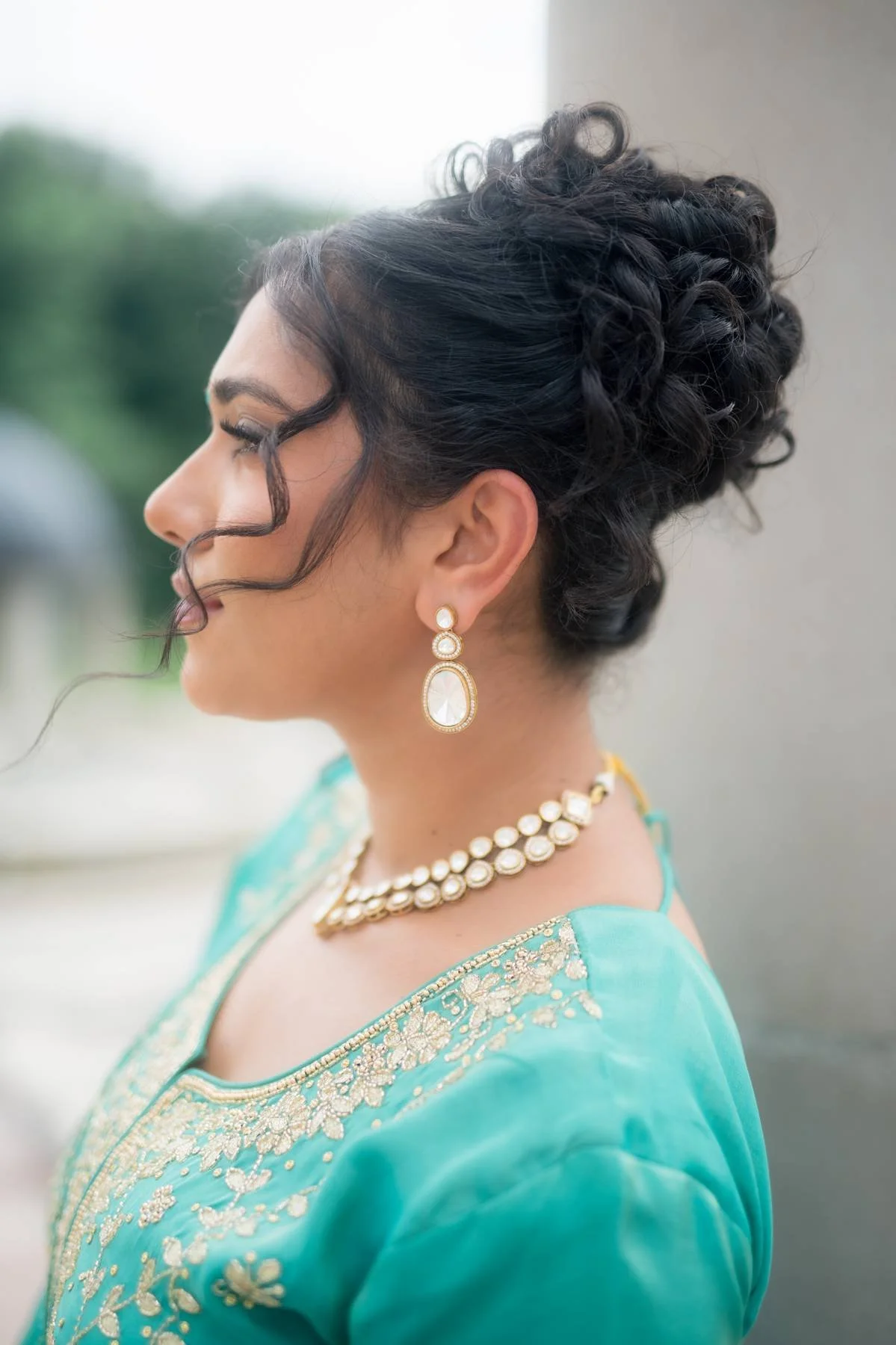Profile of a woman with dark curly hair styled in an elegant updo, wearing pearl earrings and a turquoise traditional dress with gold embroidery, outdoors on a cloudy day.