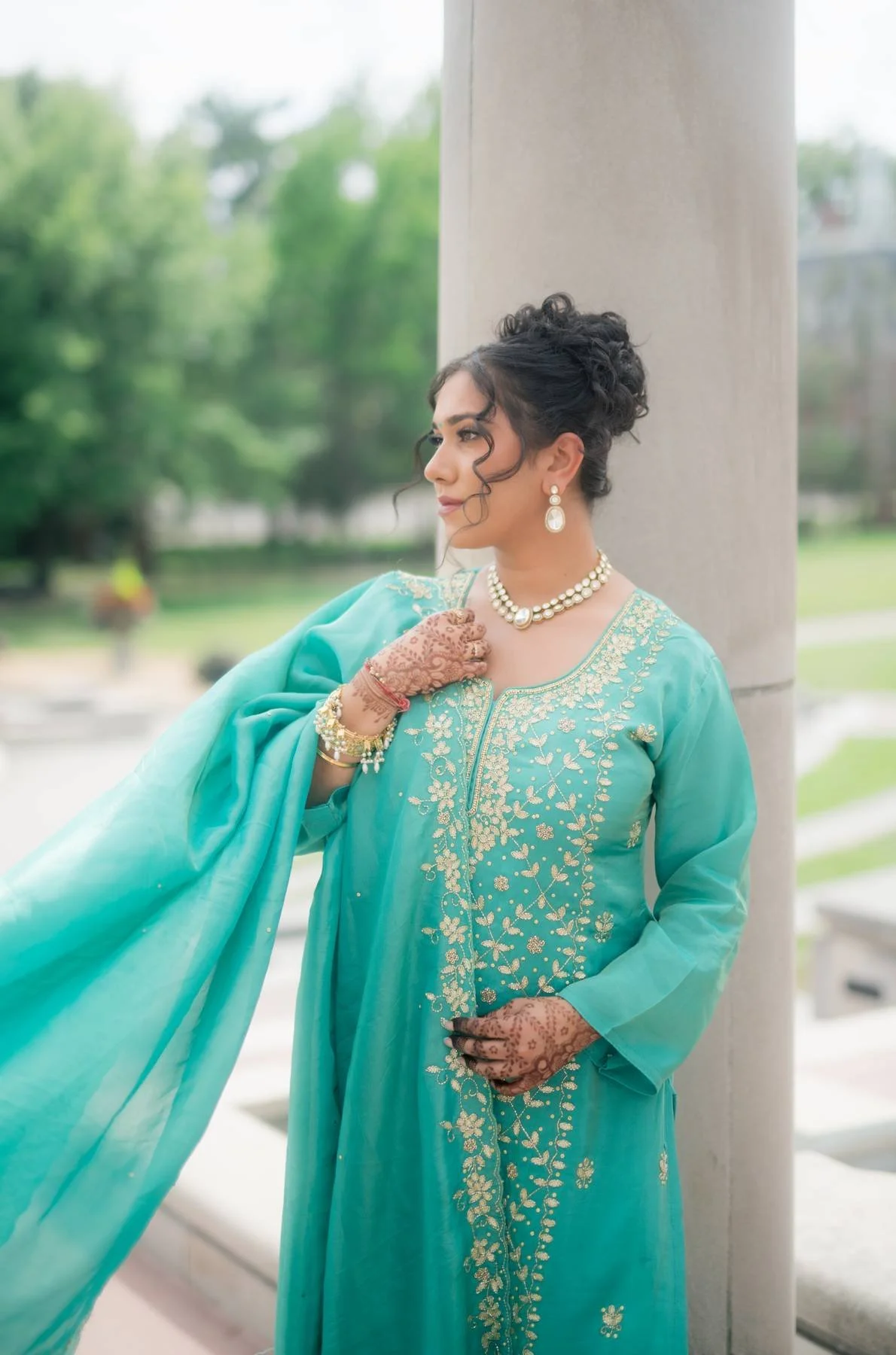 A woman in a turquoise traditional dress with gold embroidery, accessorized with pearl jewelry, standing outdoors near a large column with greenery in the background.