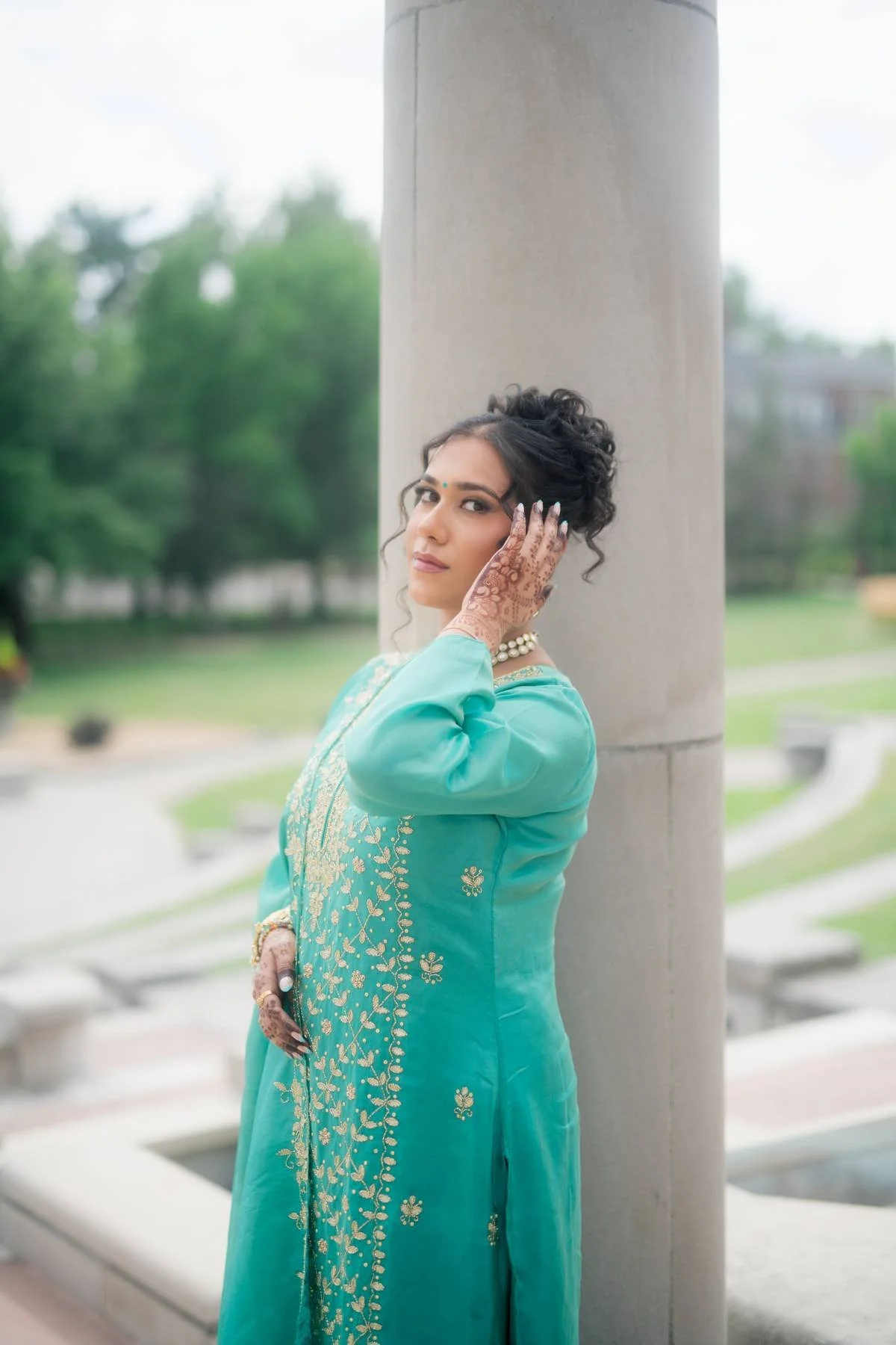 A woman in a teal and gold traditional Indian dress standing outdoors, leaning against a large concrete pillar, with henna on her hands and jewelry.