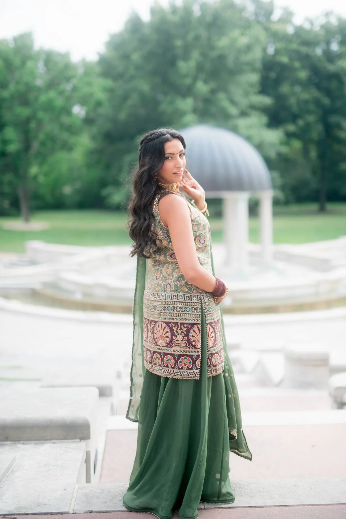 Woman in a green traditional dress with intricate embroidery posing outdoors near a fountain and trees in the background.