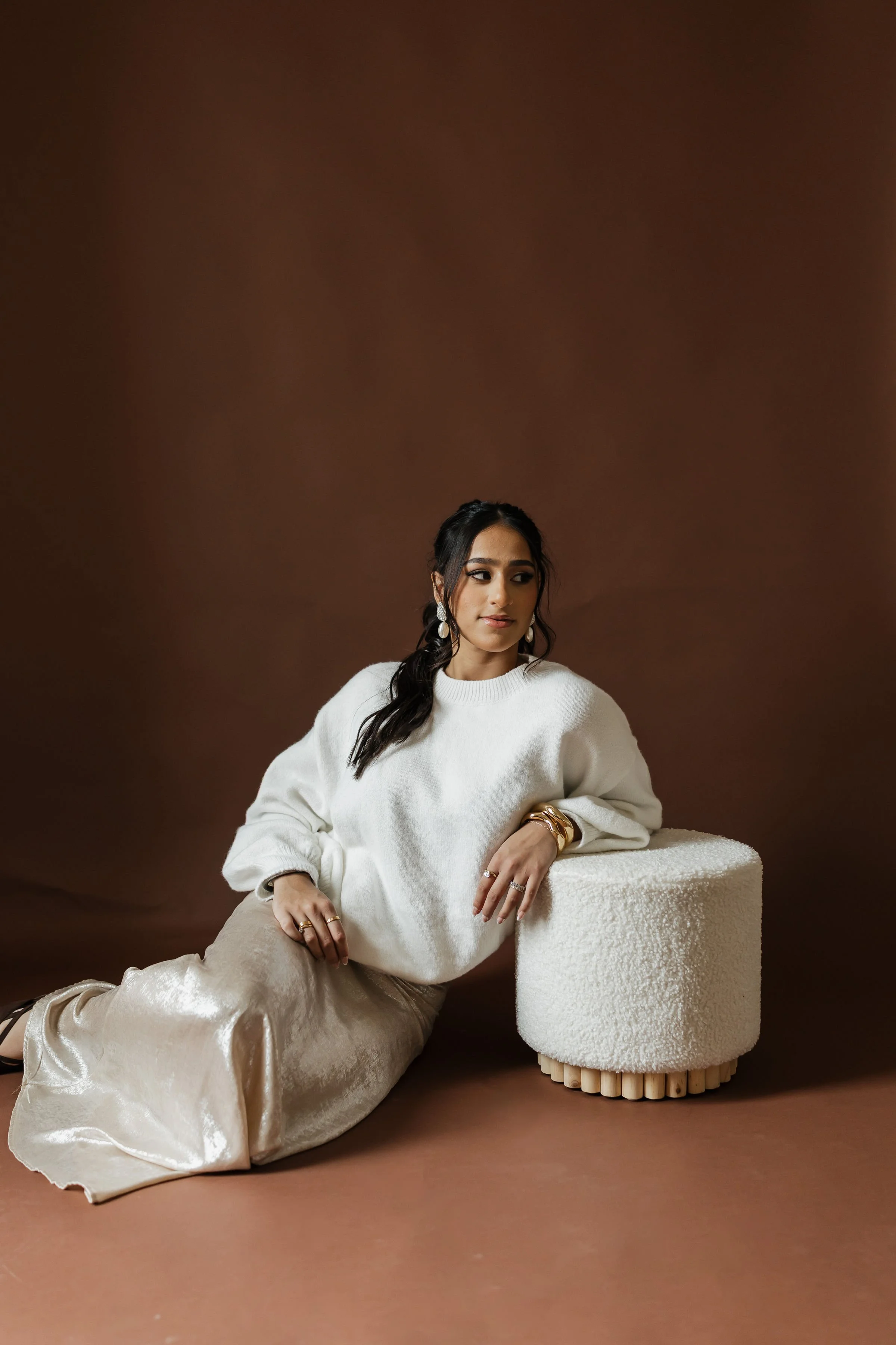 A woman with dark hair, wearing a white sweater and beige pants, sitting next to a textured white cylindrical table against a brown background, accessorized with pearl earrings, rings, and bracelets.
