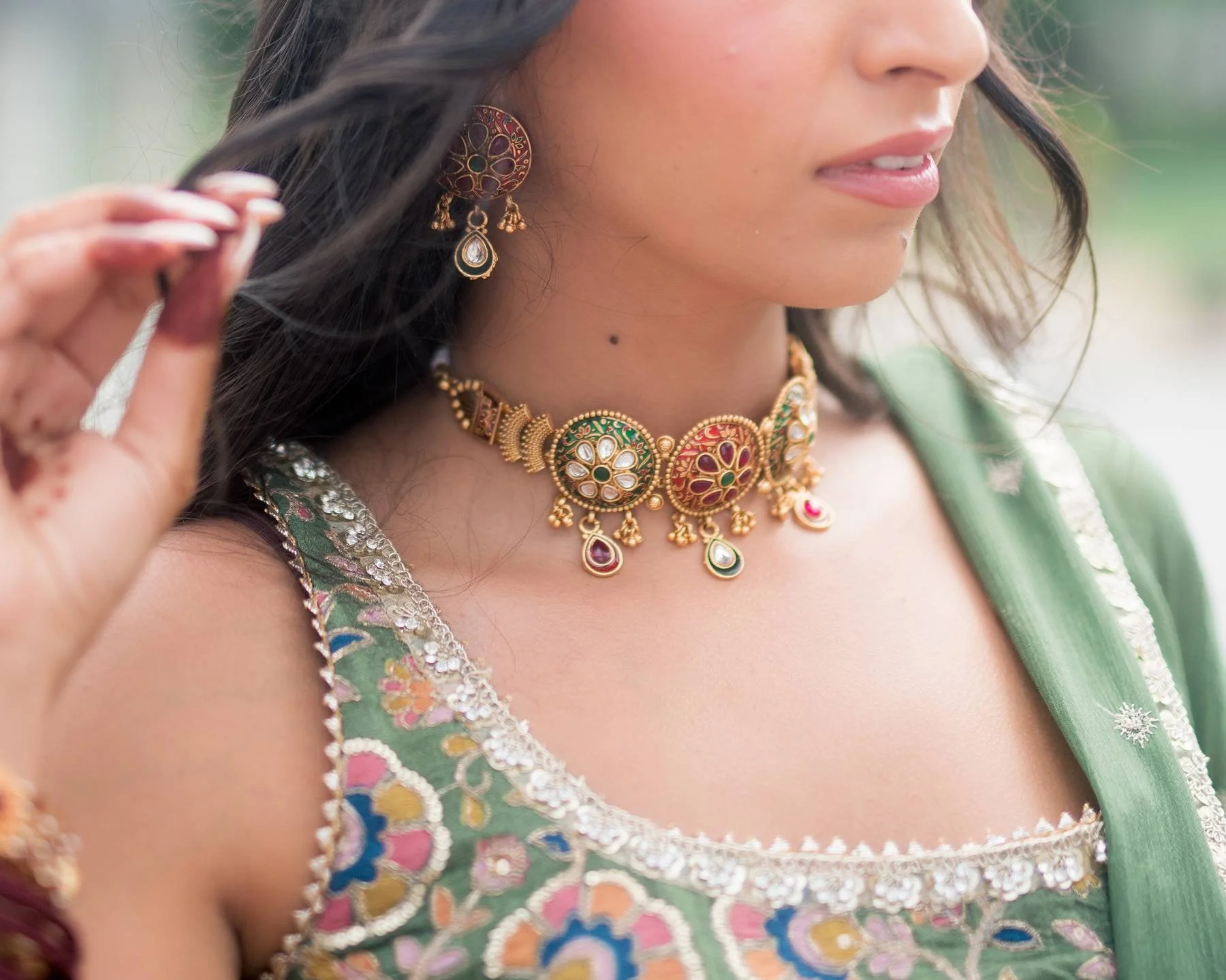 Close-up of a woman wearing traditional Indian jewelry and clothing, including a large gold necklace with colored accents, earrings, and a green saree with intricate embroidery.