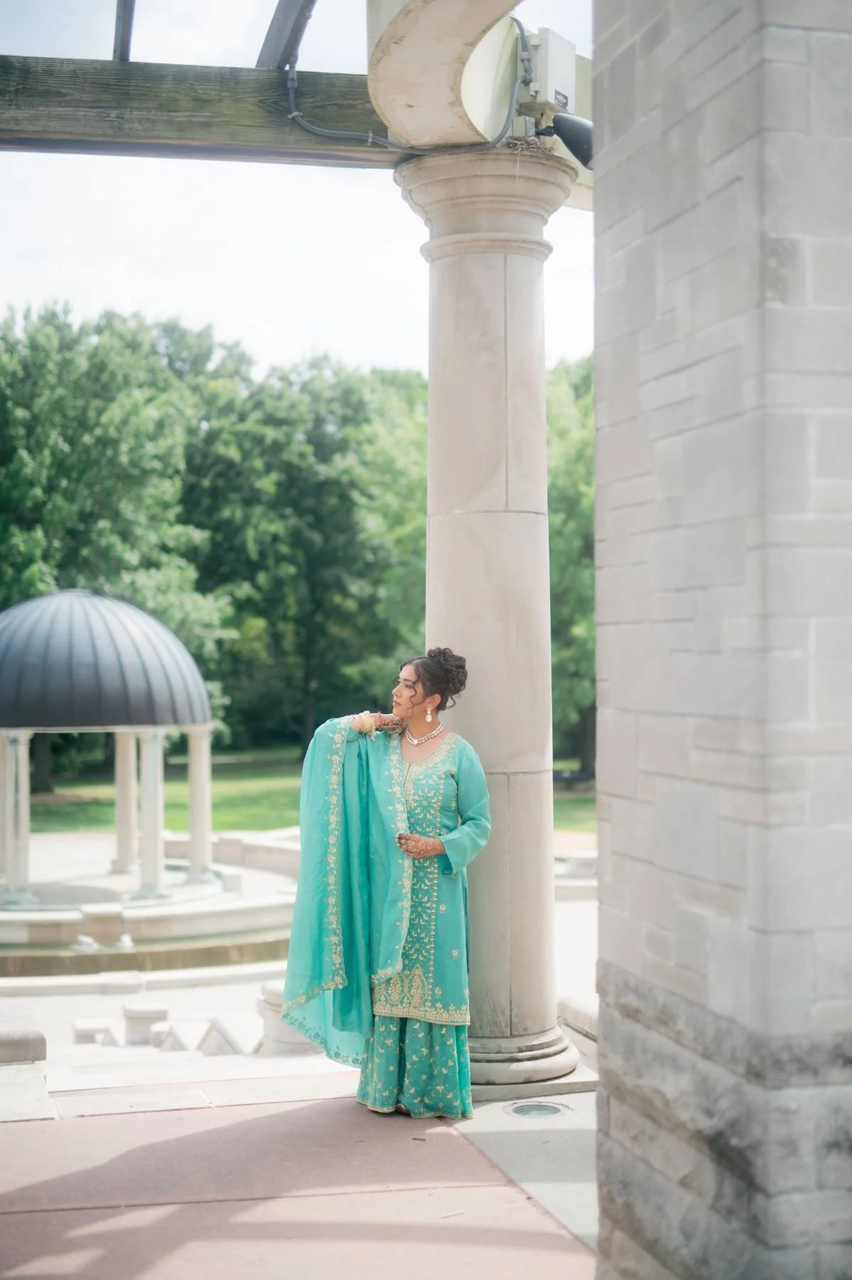 Woman in traditional turquoise Indian attire standing near a large stone column and decorative fountain, with a green park background.