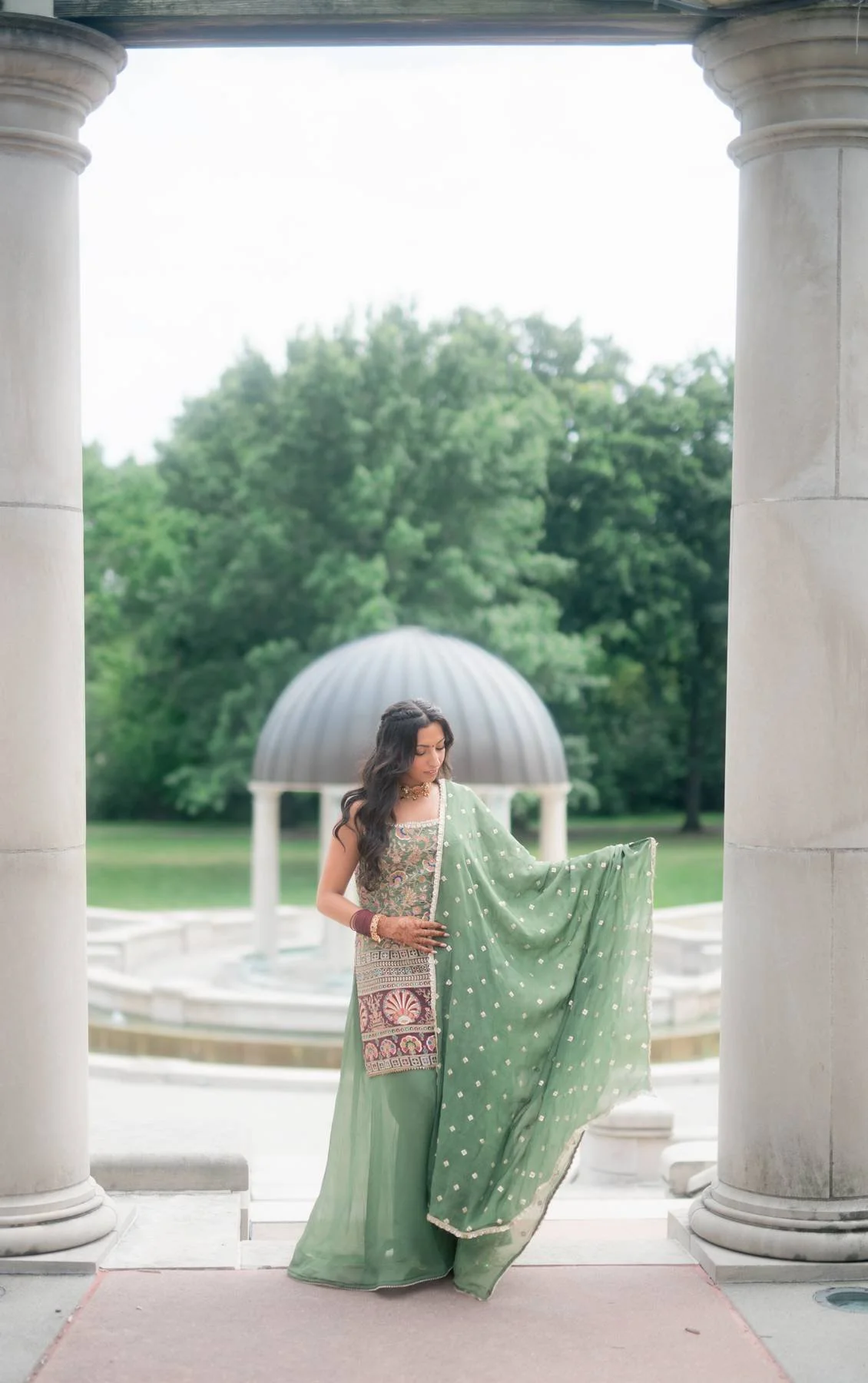 A woman in traditional Indian attire standing between marble columns, holding a green dupatta with her left hand, with a fountain and lush green trees in the background.