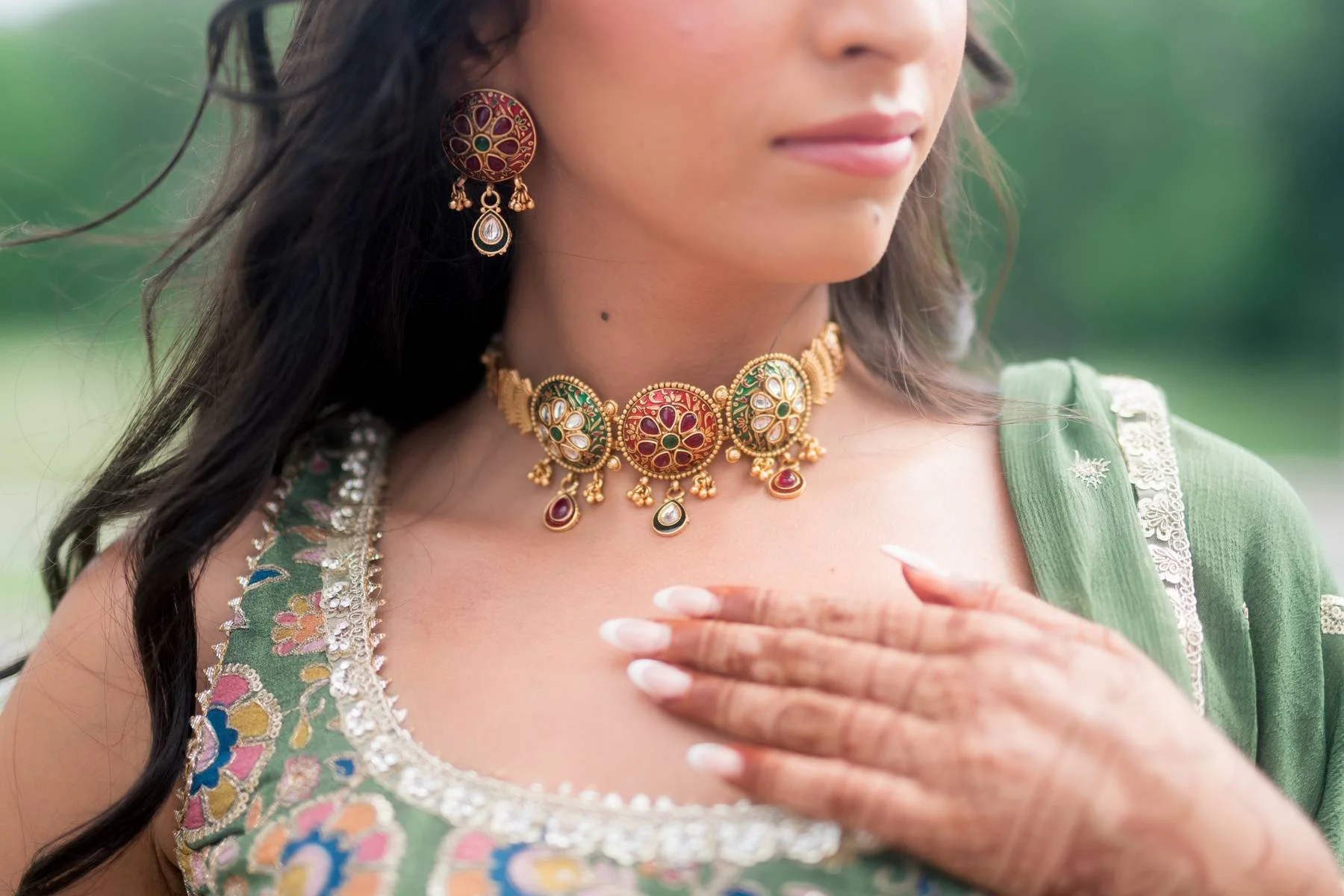 Close-up of a woman wearing traditional Indian jewelry and a green embroidered outfit. She has a hand on her chest and is outdoors with a blurred green background.