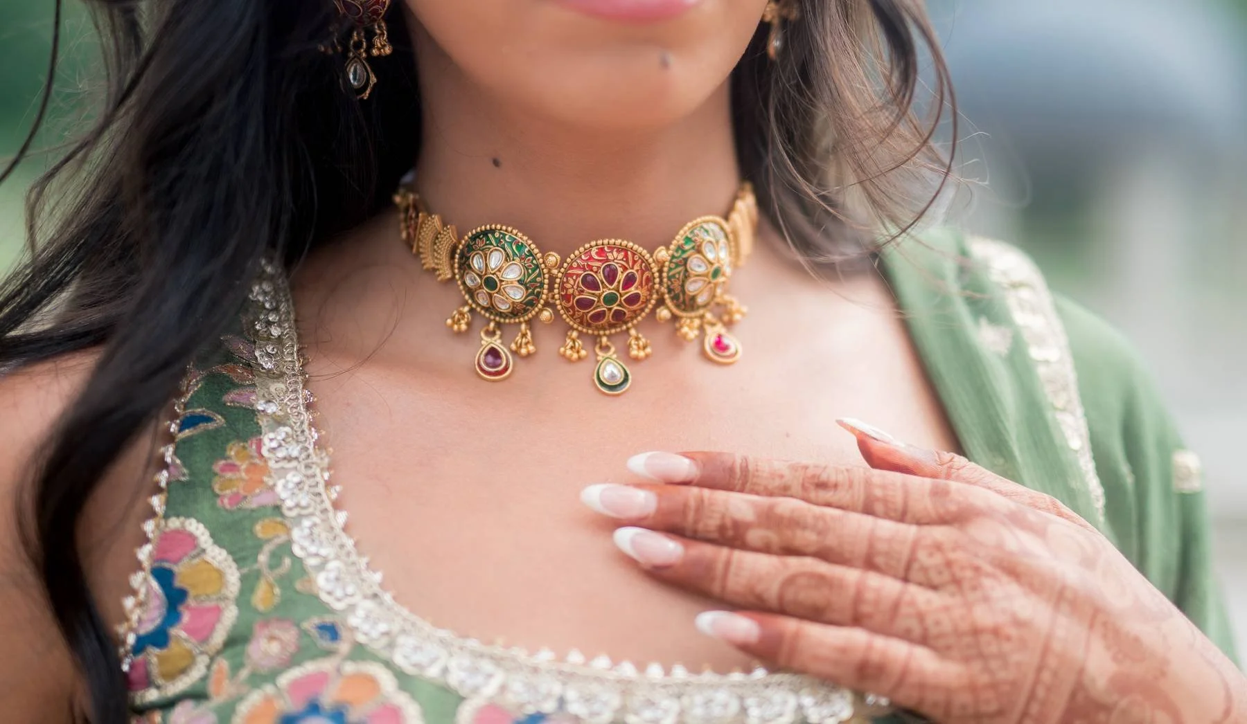 Close-up of a woman wearing intricate jewelry, including a gold necklace with colorful stones and dangling embellishments, and a green traditional outfit with colorful embroidery.