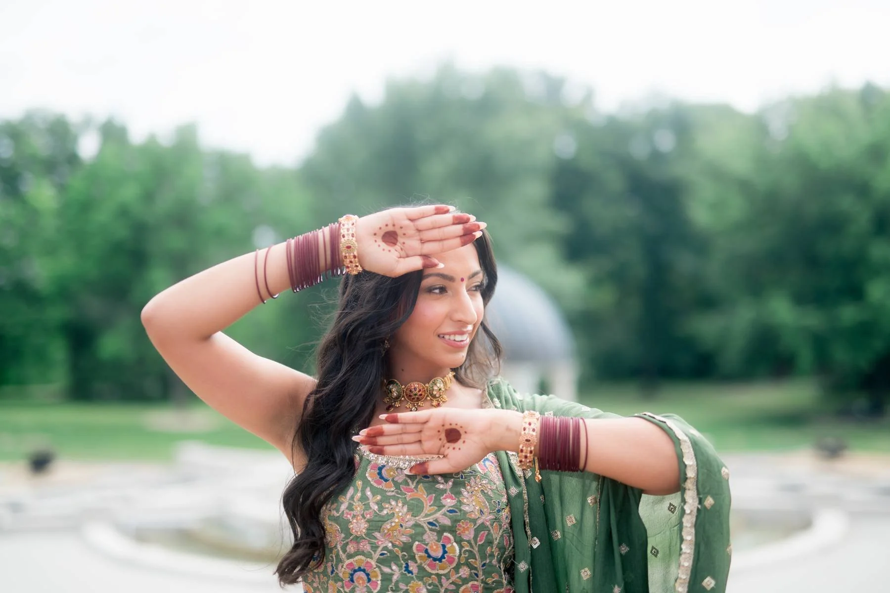 A woman dressed in traditional Indian attire performing a dance pose outdoors, with her hands positioned near her face and waist, wearing jewelry and bangles, with a blurred green park background.