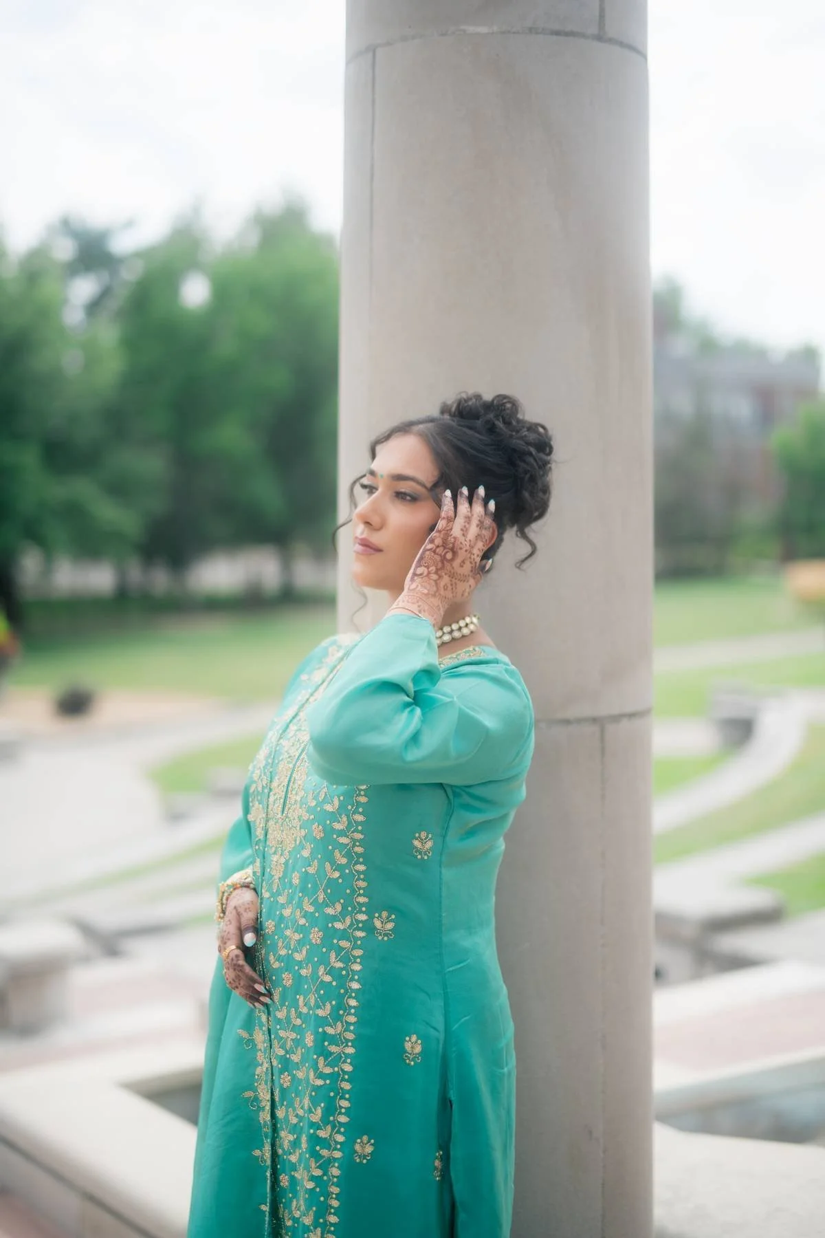 A woman in a teal traditional dress, standing outdoors, leaning against a large concrete pillar. She has intricate henna on her hands and is wearing jewelry, including a necklace and bangles, with her hair styled in curls.