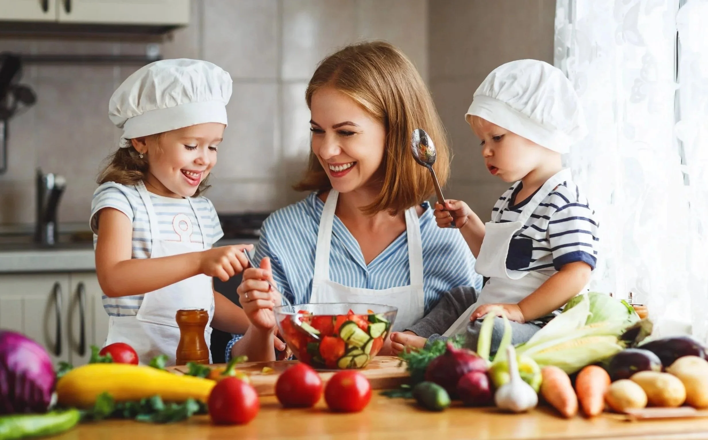 A woman and two young girls wearing chef hats and aprons preparing food in a kitchen with fresh vegetables on the table.