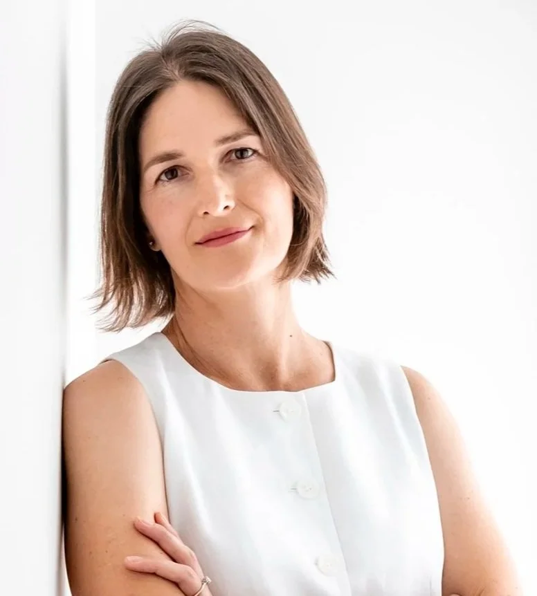 Portrait of a Jennifer, a woman with short brown hair, wearing a sleeveless white top, smiling softly, standing against a white background.