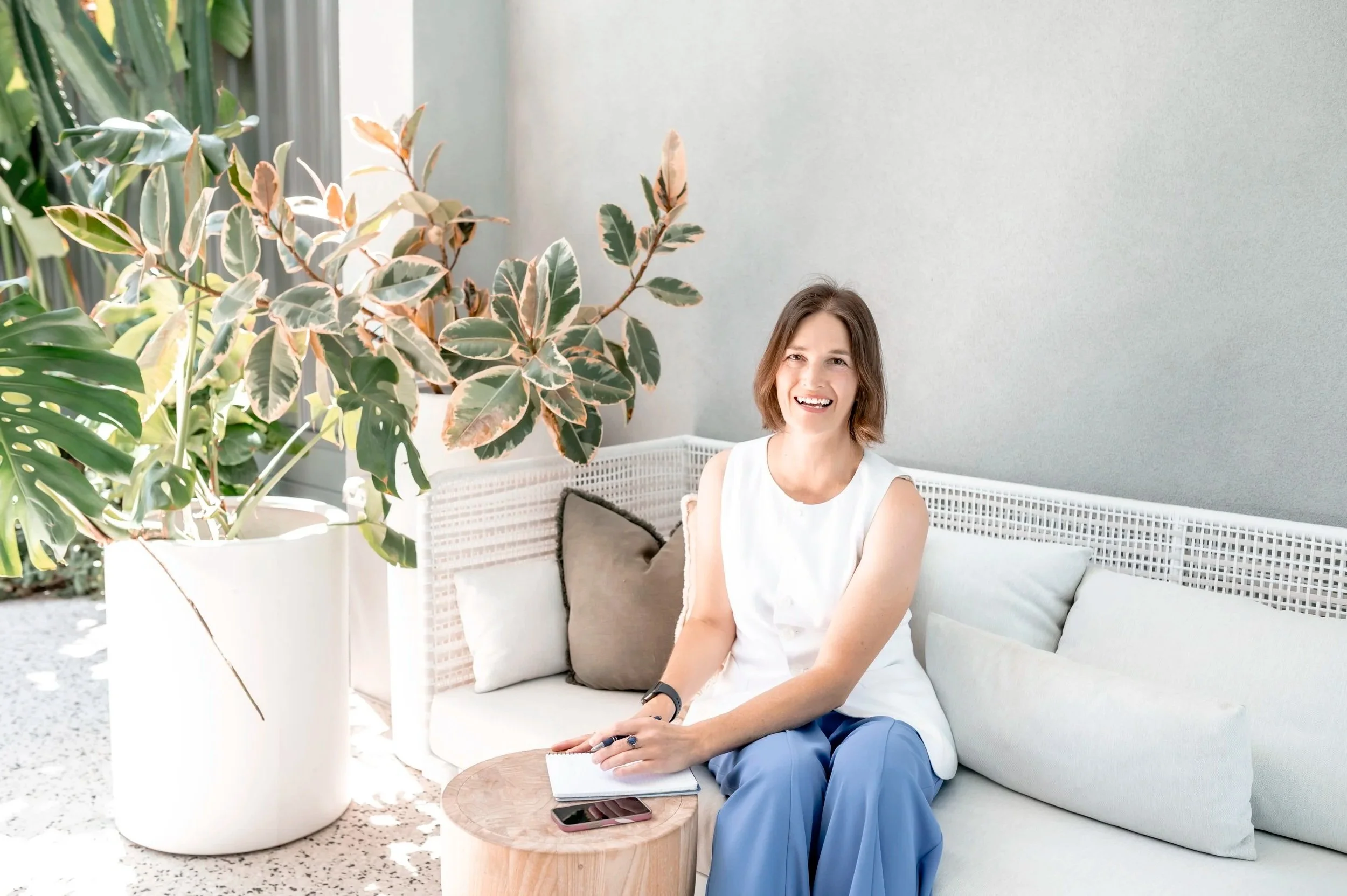 A woman with shoulder-length brown hair smiling and sitting on a white outdoor sofa with beige cushions. She is wearing a white sleeveless top and blue pants, with a notebook and a pink smartphone on a small round wooden table in front of her. There are large green and variegated leaves from a tall plant in a white pot to her left. The background features a light grey wall and sunlight filtering through the greenery.