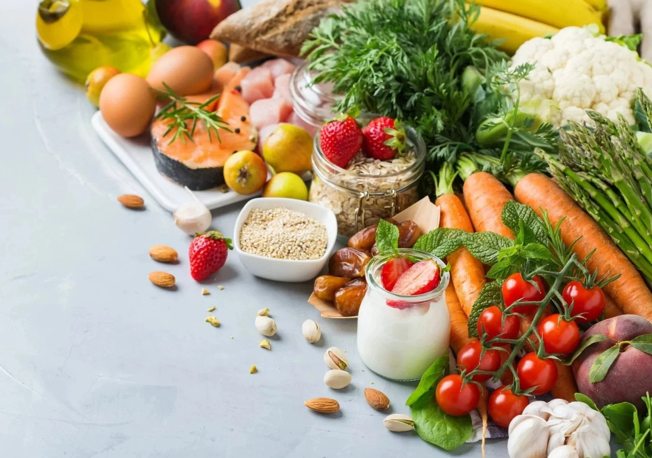 Fresh vegetables, fruits, eggs, grains, and herbs arranged on a light-colored table, including carrots, cherry tomatoes, cauliflower, strawberries, apples, and parsley.