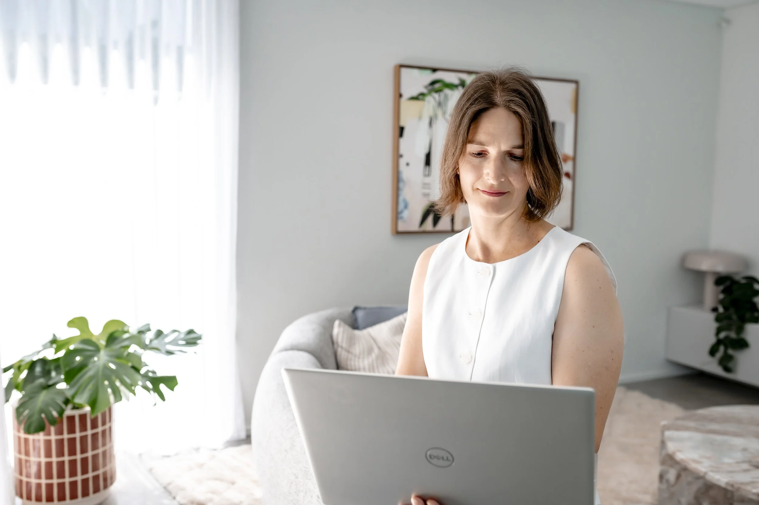 A woman with shoulder-length brown hair, wearing a white sleeveless blouse, working on her laptop in a bright, modern room. There is a plant in a striped pot on the table, a window with sheer curtains, and artwork hanging on the wall behind her.