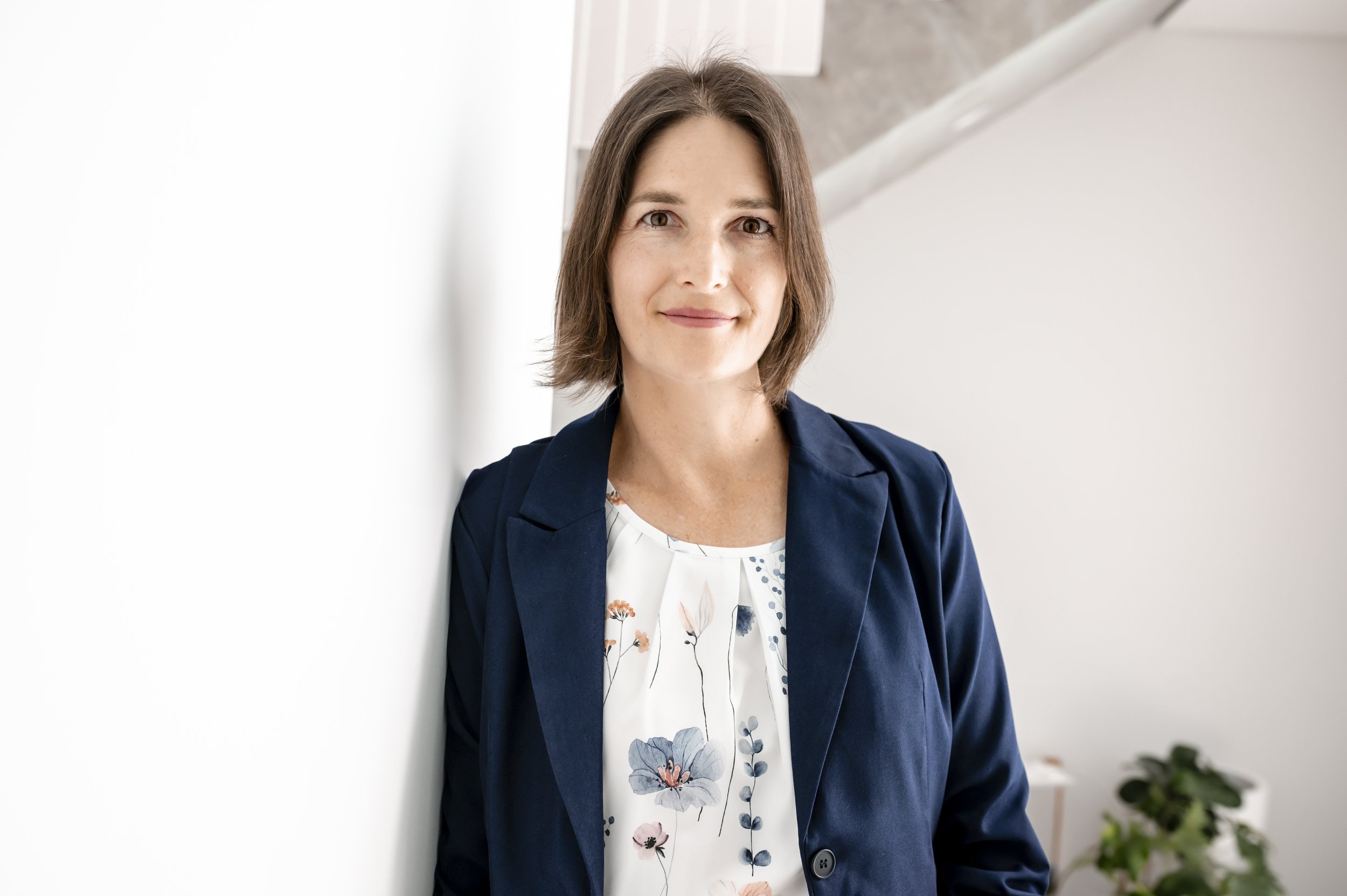 Jennifer - A woman with shoulder-length brown hair, wearing a navy blazer and a white floral blouse, standing indoors next to a white wall with a staircase in the background