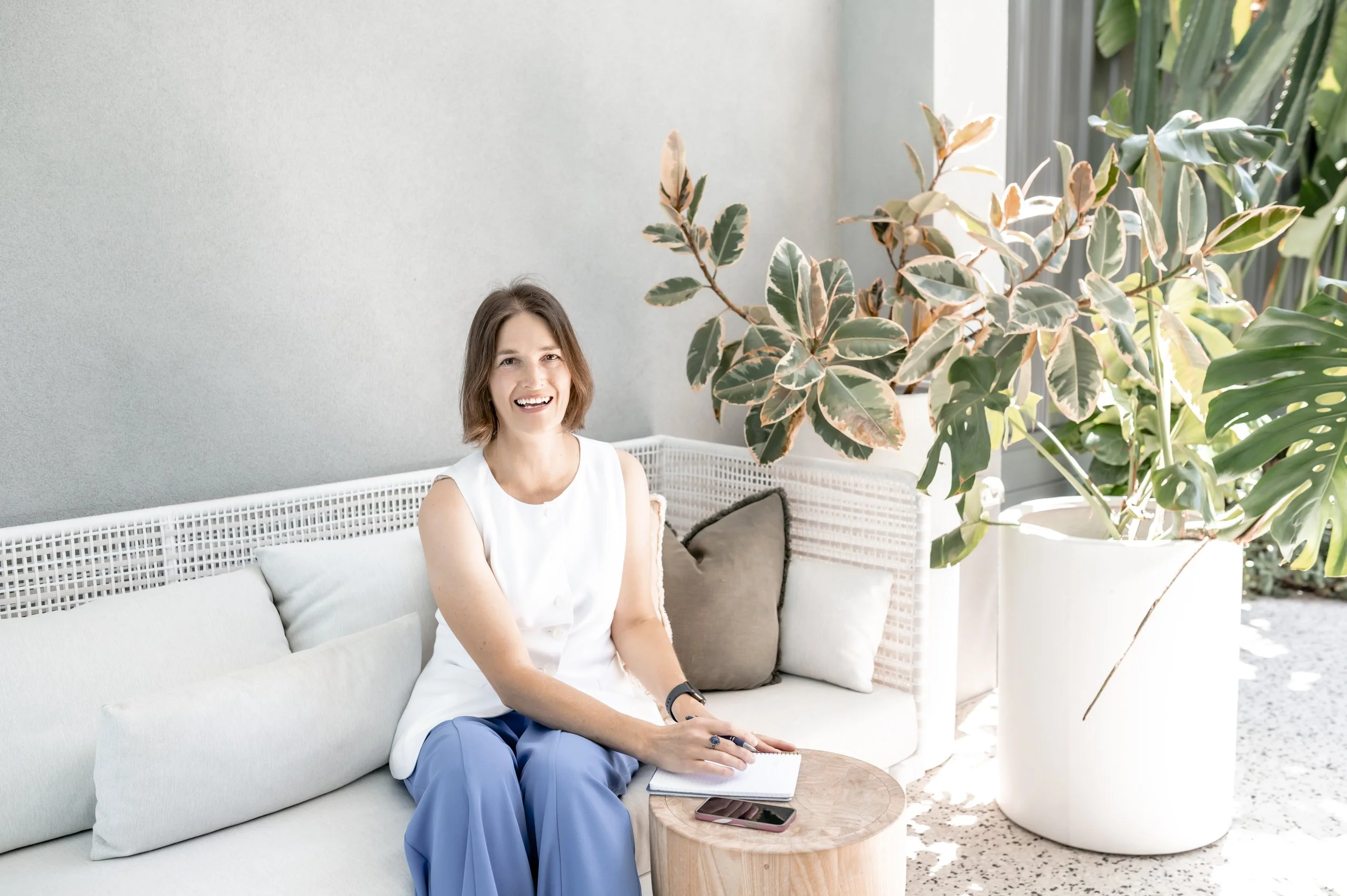A smiling woman with short brown hair wearing a white sleeveless top sitting on a light-colored outdoor sofa next to a wooden side table, a smartphone and notebook on her lap, surrounded by large indoor/outdoor plants in a bright setting with a gray wall behind her.