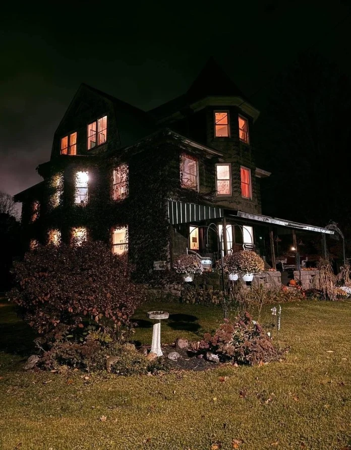 A large, dark Victorian house at night with warm lights glowing from the windows. The house has a porch with potted plants and a garden with bushes and flowers in the front yard.