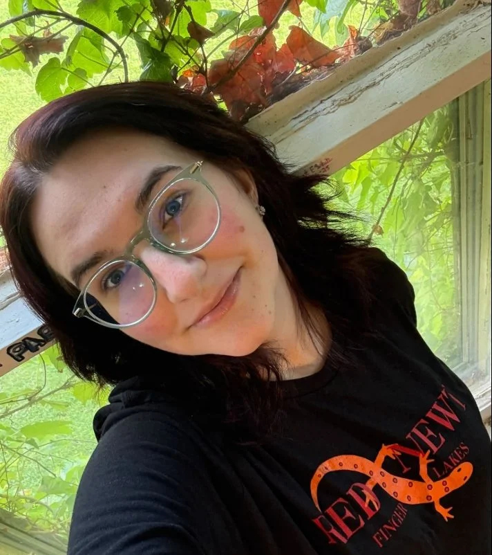 A woman with dark hair and glasses taking a selfie in front of a window with green and red plant leaves outside.