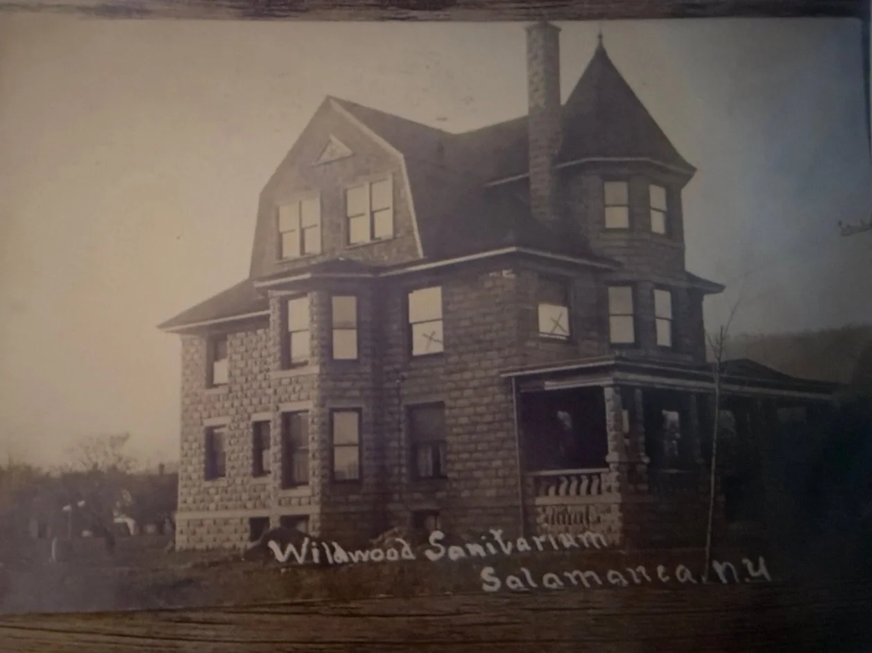 An old photograph of the Wildwood Sanitarium located in Salamanca, New York, featuring a large, three-story Victorian-style brick building with multiple gabled roofs, a tower, and numerous windows with some marked with an 'X'.