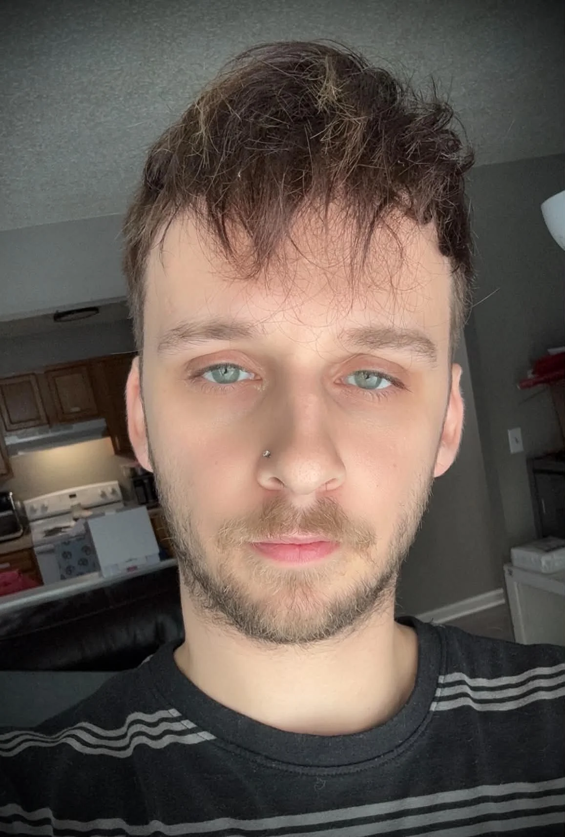 Close-up of a young man with light skin, blue eyes, and facial hair, wearing a black and gray striped shirt, in a home interior with kitchen cabinets and appliances in the background.