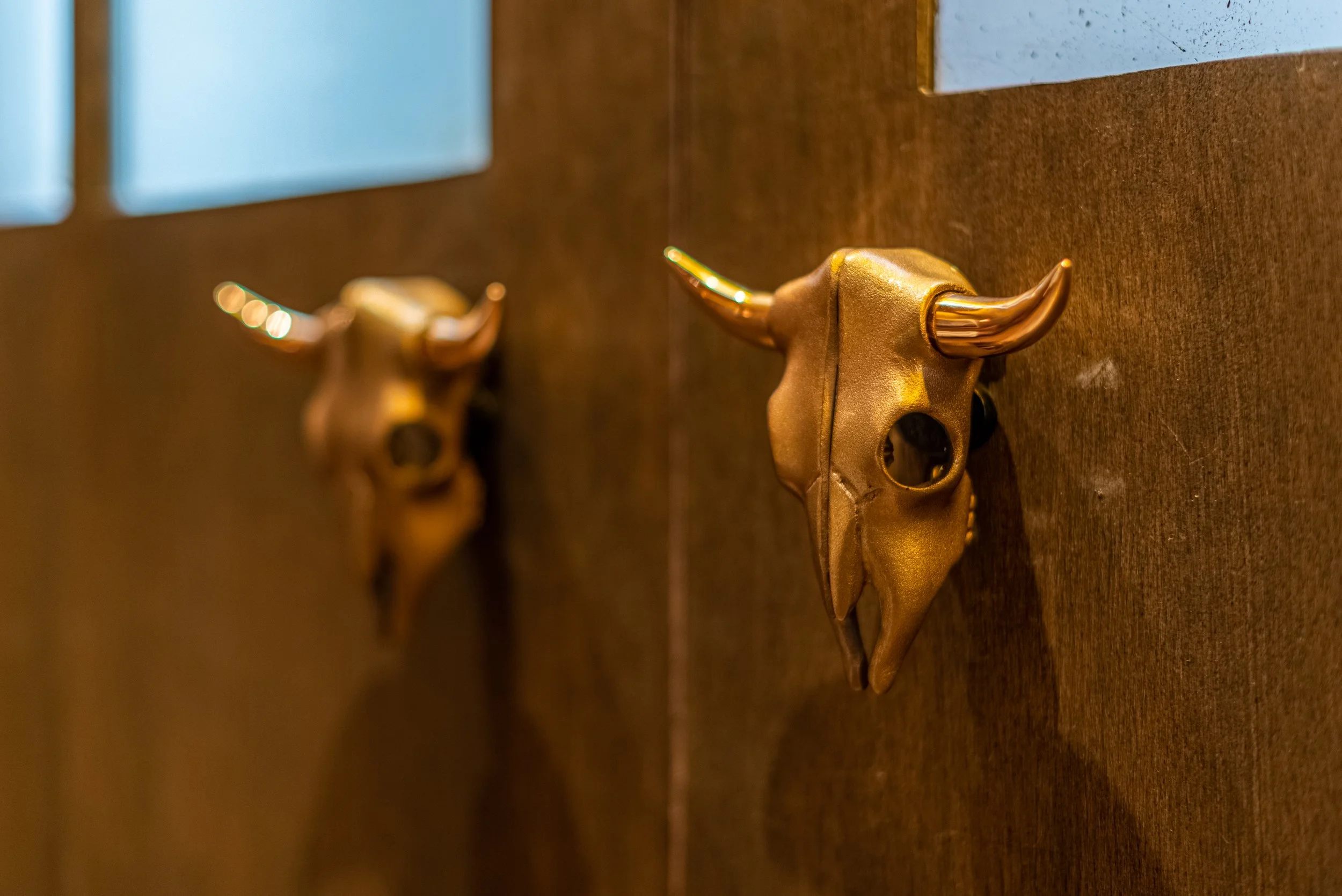 Decorative wall-mounted bull skull with metallic horns and gold finish, reflected in a mirror, against a brown textured wall.