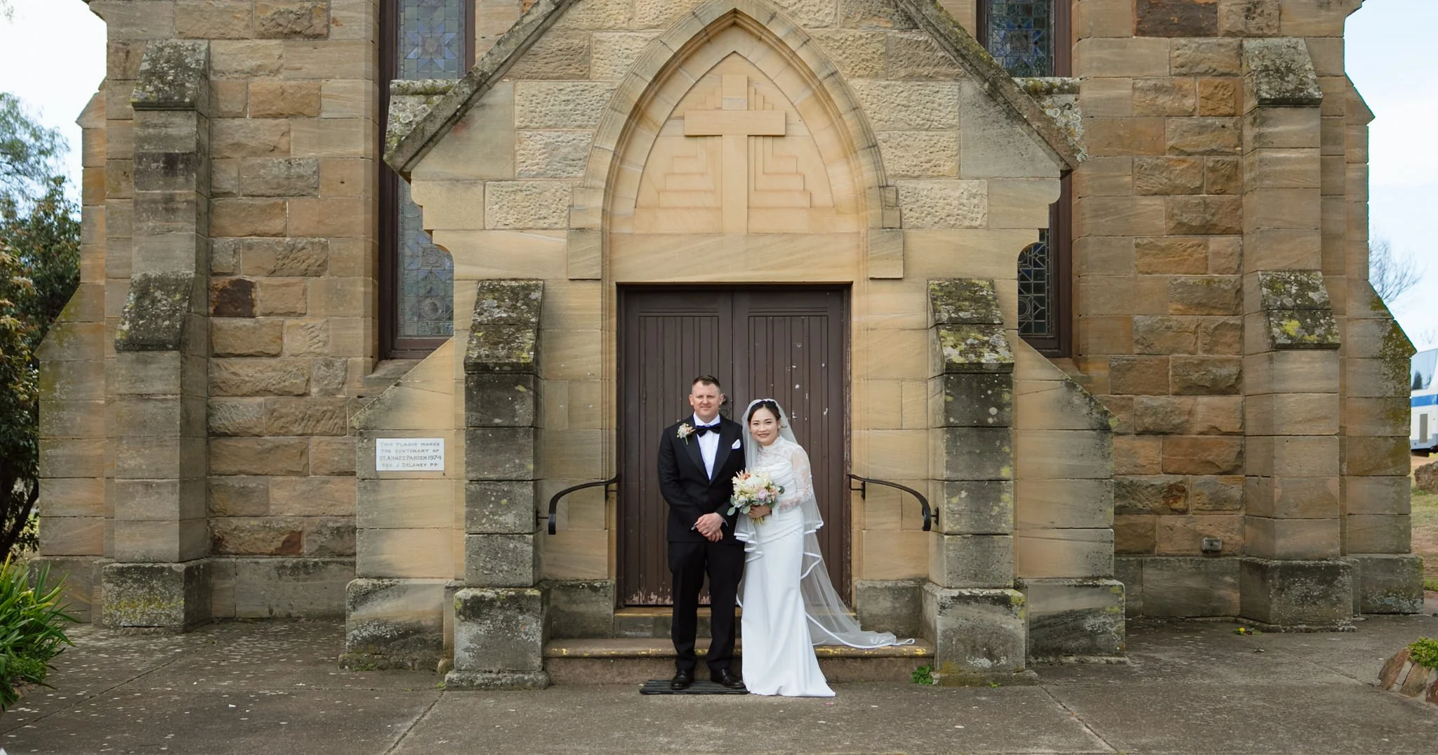 Bride and groom standing together outside the sandstone entrance of Merriwa Catholic Church.
