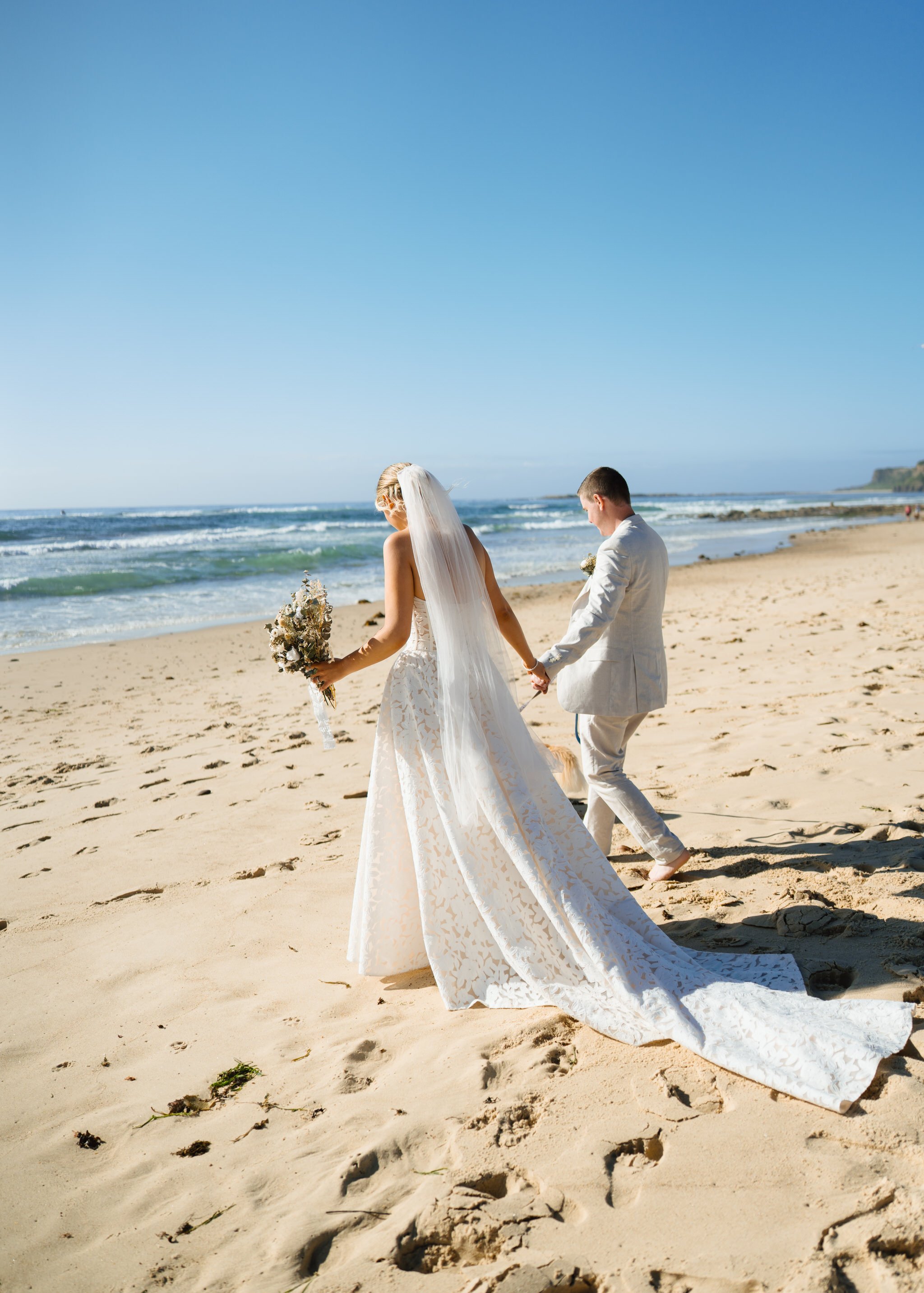 Bride and groom walking barefoot along shoreline holding hands at Caves Beach