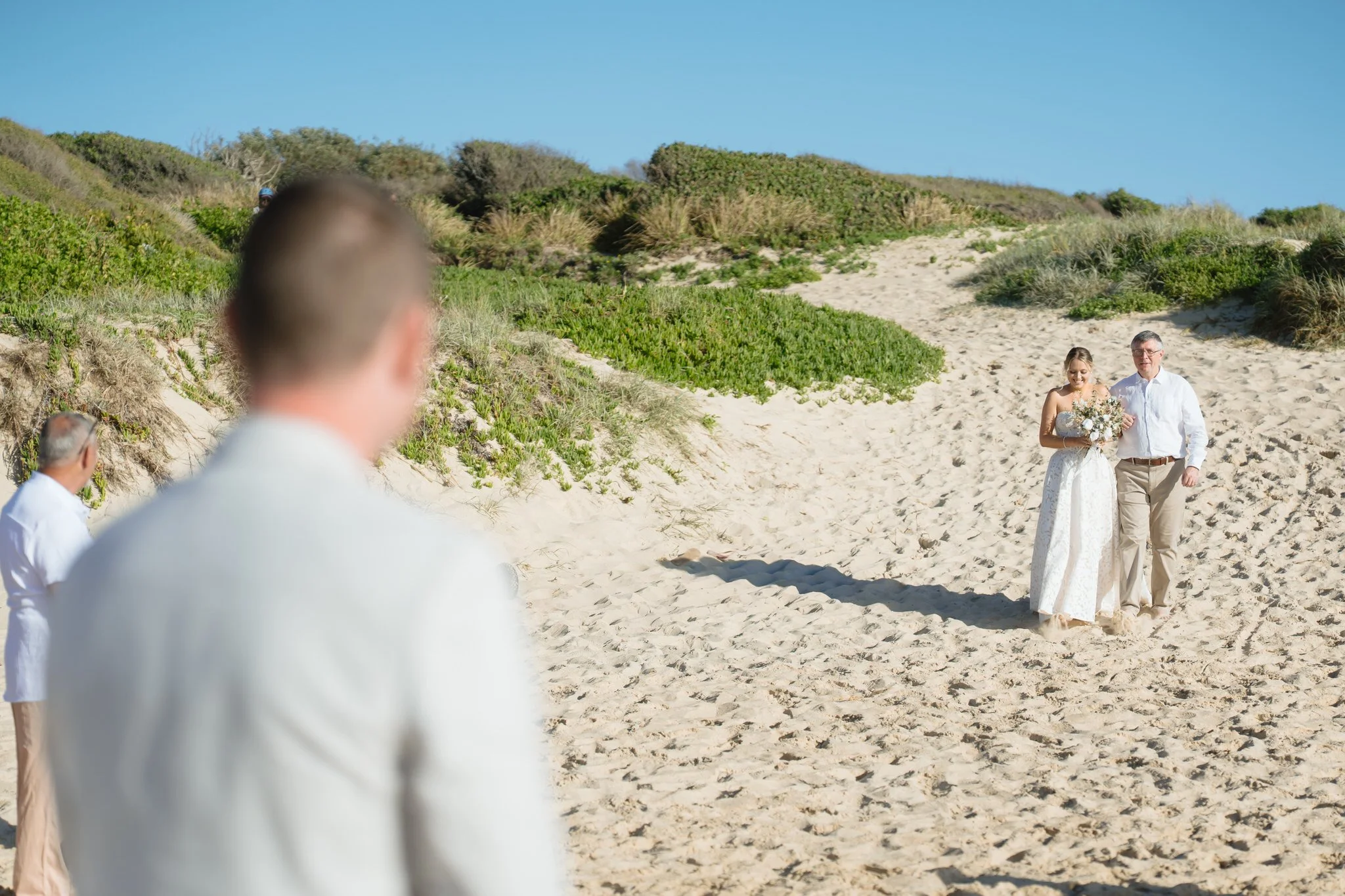 Bride walking down sandy path with her father before beach ceremony at Caves Beach