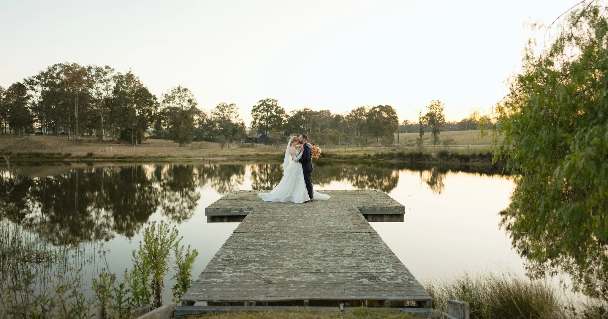 Bride and groom embracing at the end of a wooden jetty, with trees reflected in the water at Squire's Vineyard.