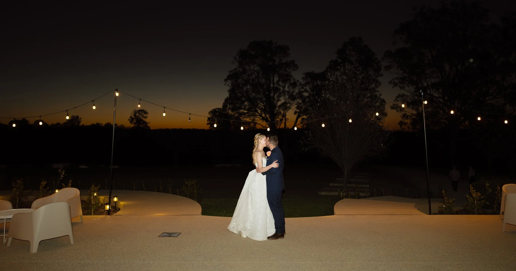 Newlyweds kissing beneath festoon lights on the outdoor terrace at Squire's Vineyard as the sky darkens.