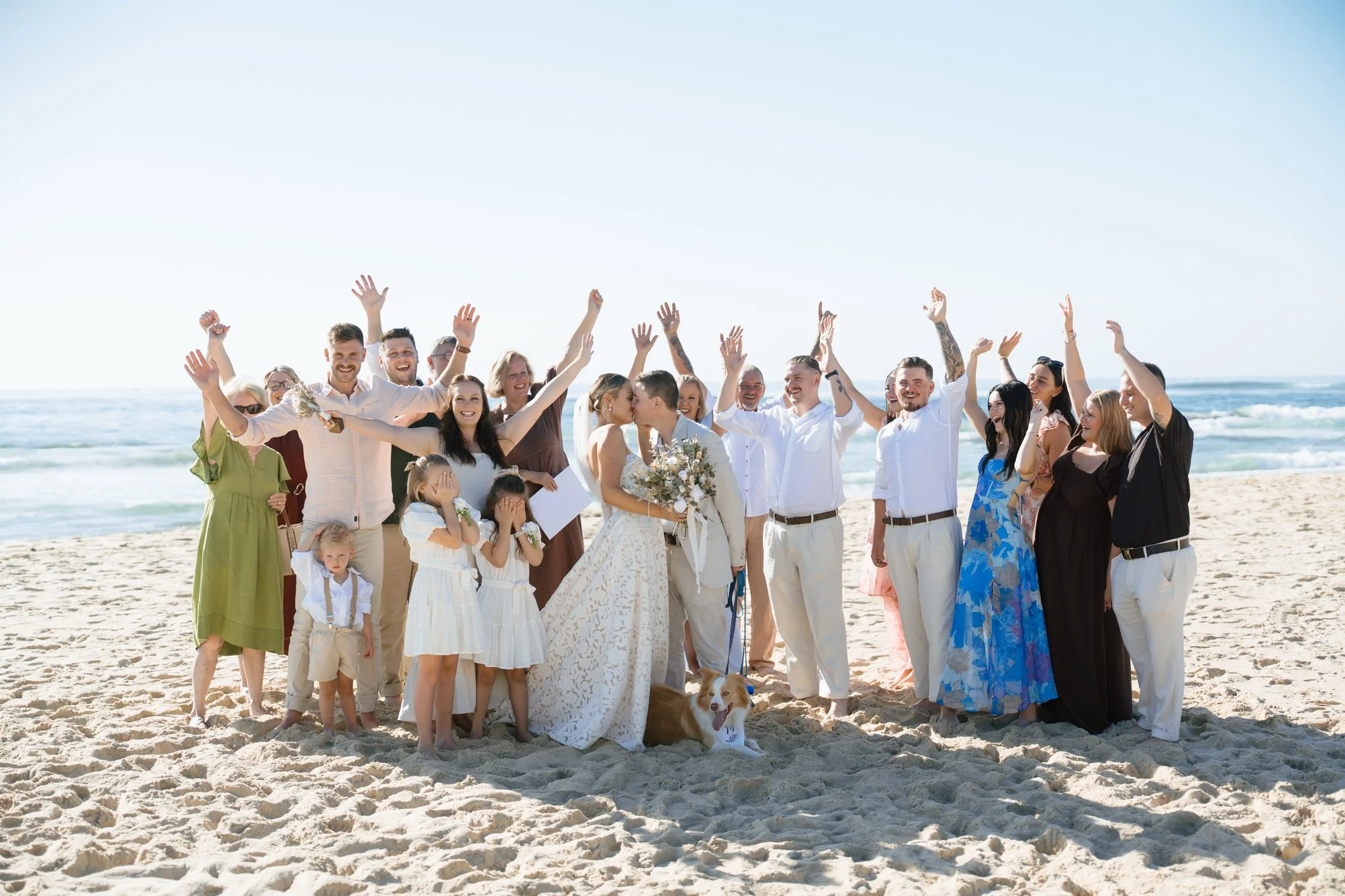 Wedding guests cheering together on the beach with couple and dog at Caves Beach
