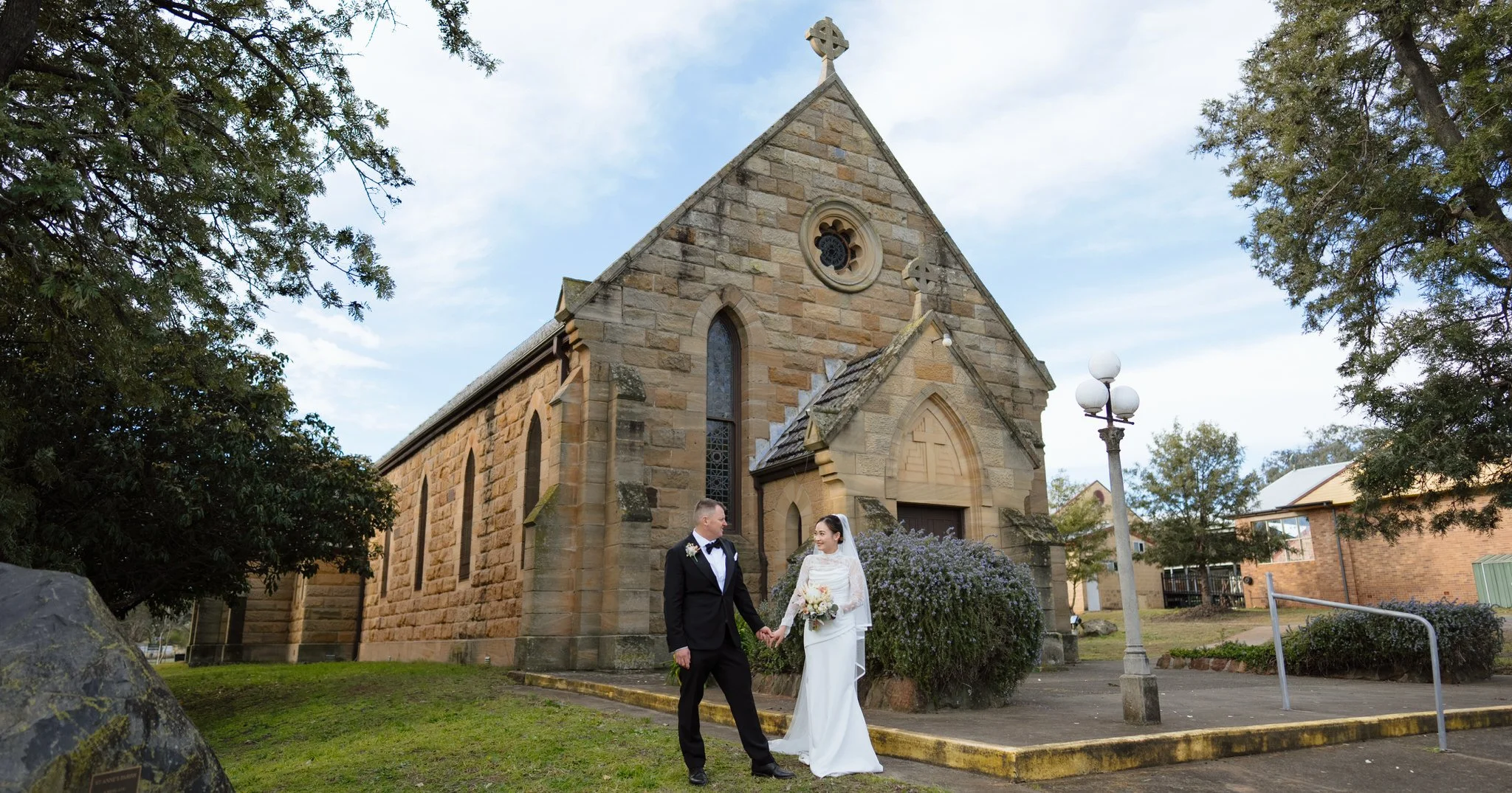 Bride and groom standing together beside the historic sandstone Merriwa Catholic Church.