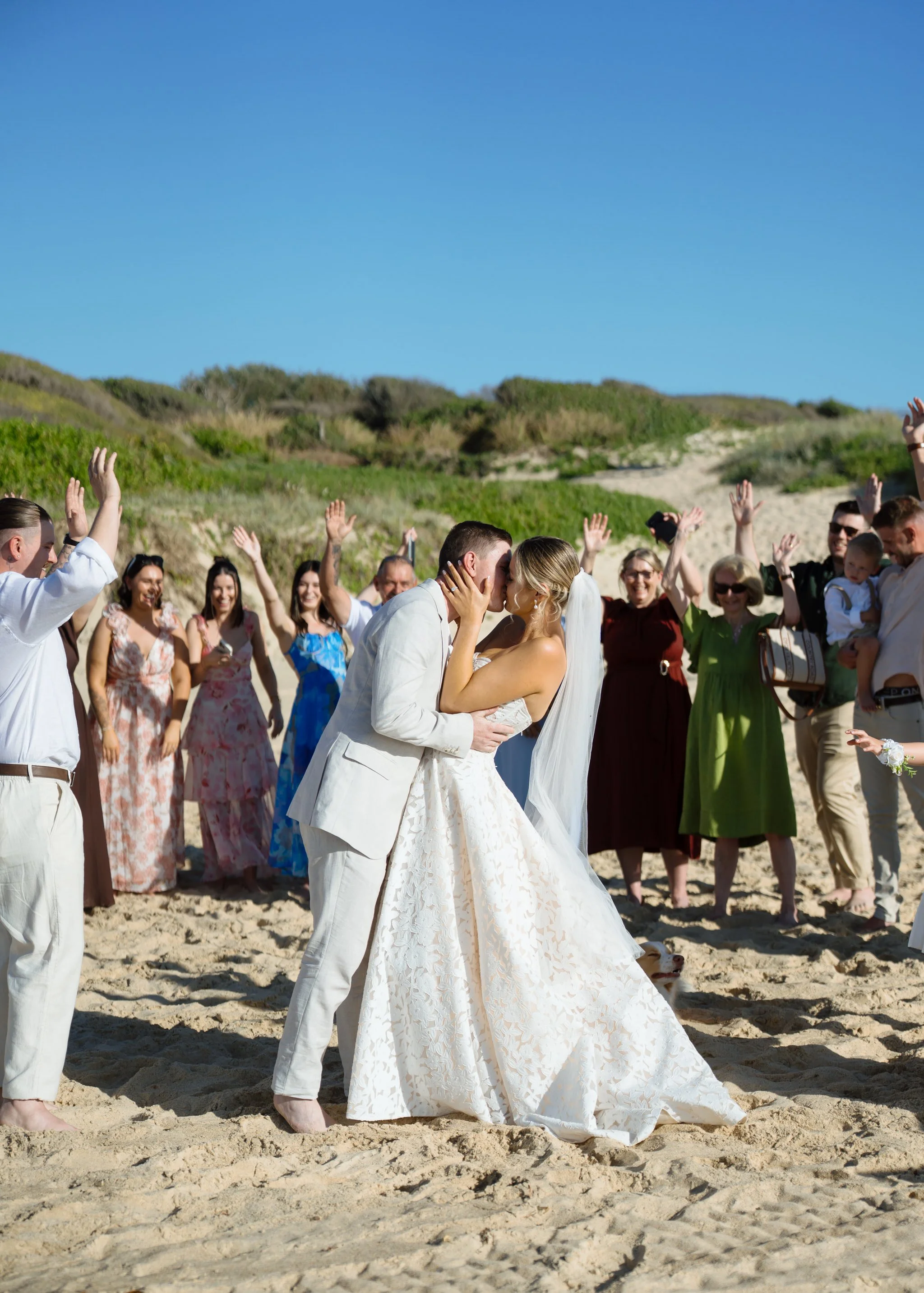 Couple sharing first kiss as guests cheer around them on the sand at Caves Beach