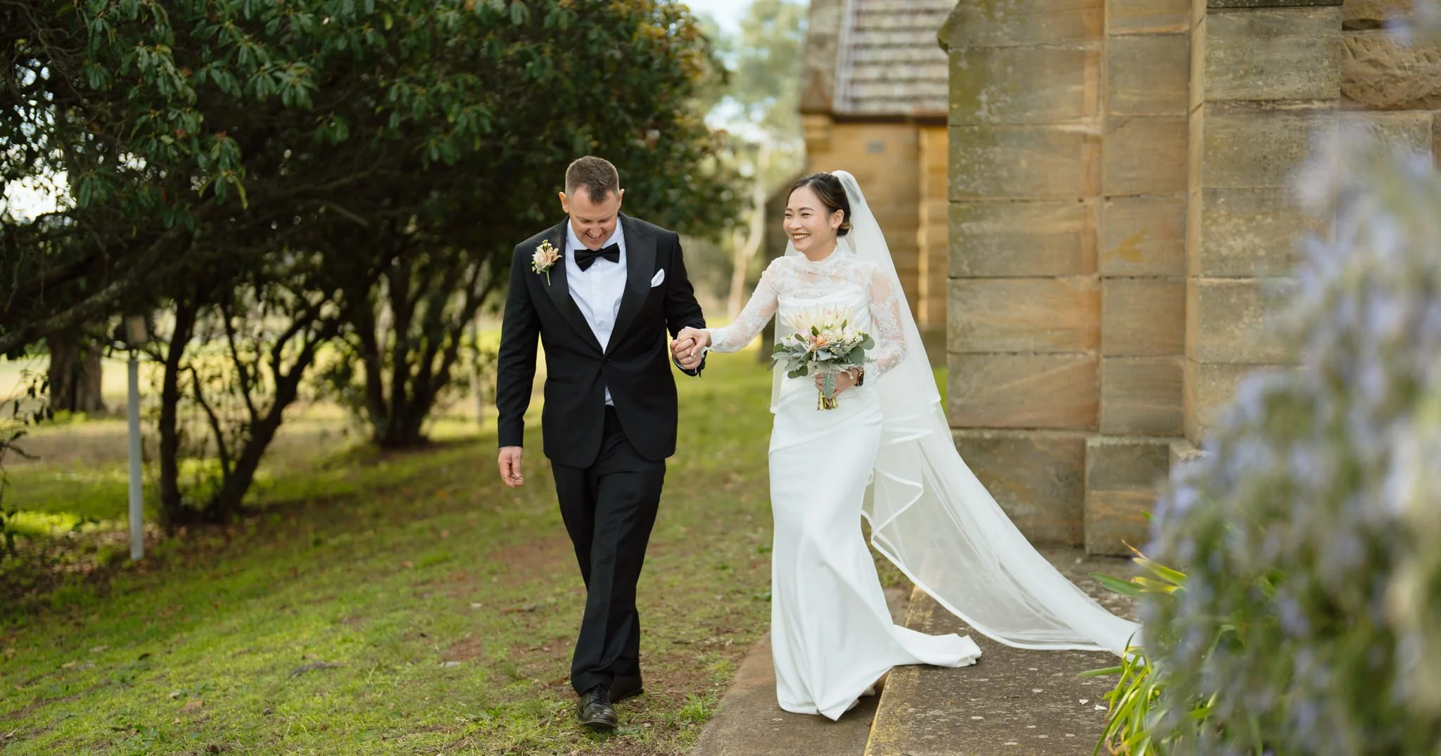 Newly married couple smiling and walking hand in hand beside Merriwa Catholic Church.