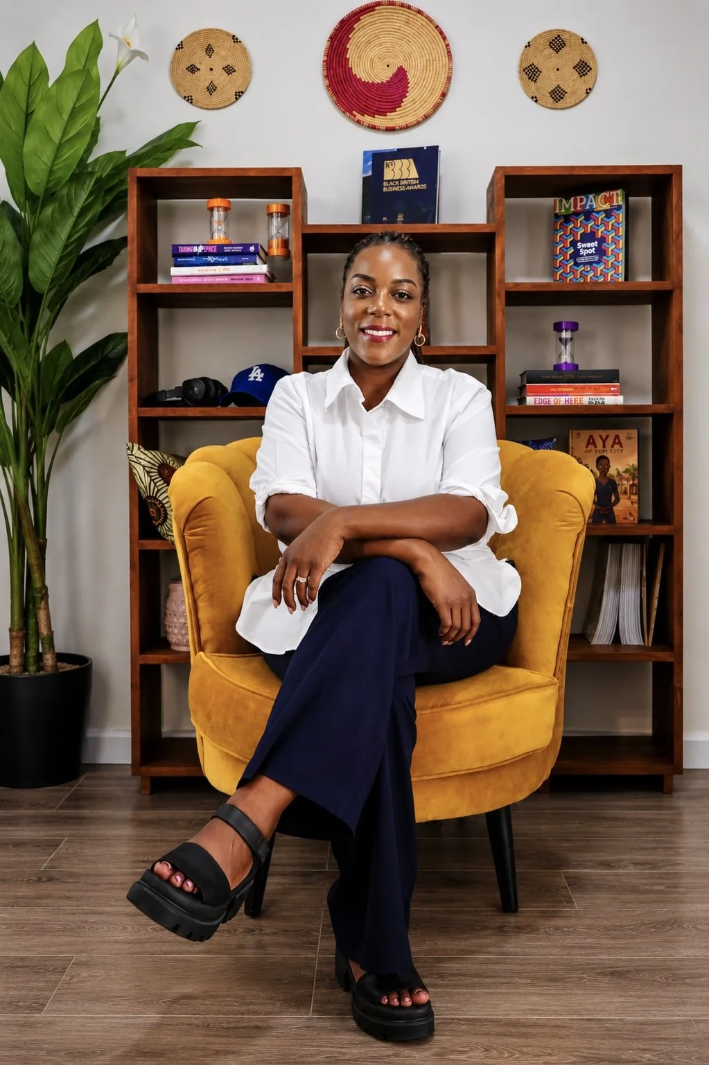 Woman sitting in a mustard yellow armchair in front of wooden shelves with books and decorative items, including a small Los Angeles Dodgers cap, plant, and wall art.