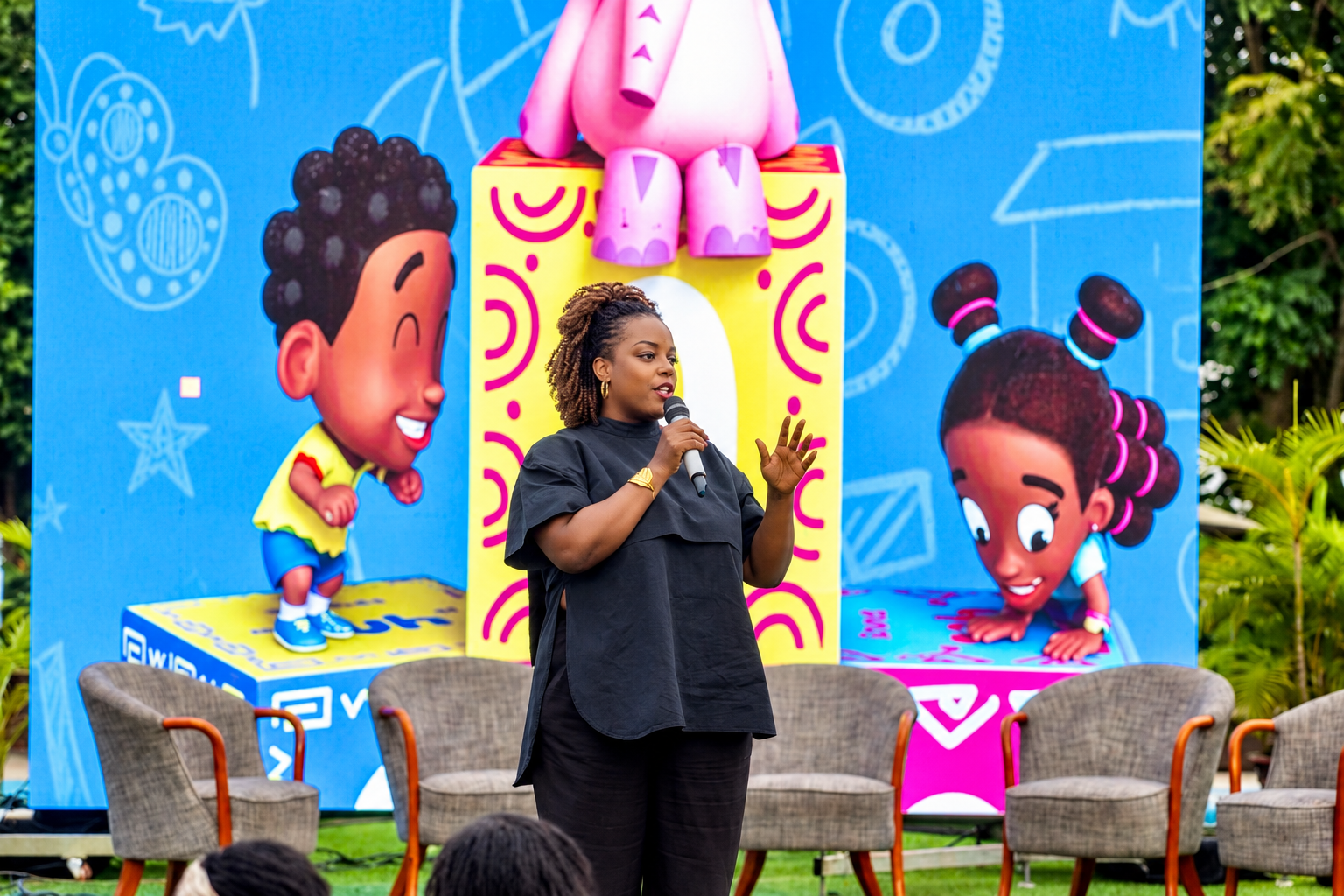 A woman standing on a stage holding a microphone, speaking at an outdoor event, with a colorful cartoon backdrop featuring diverse children and playful elements.