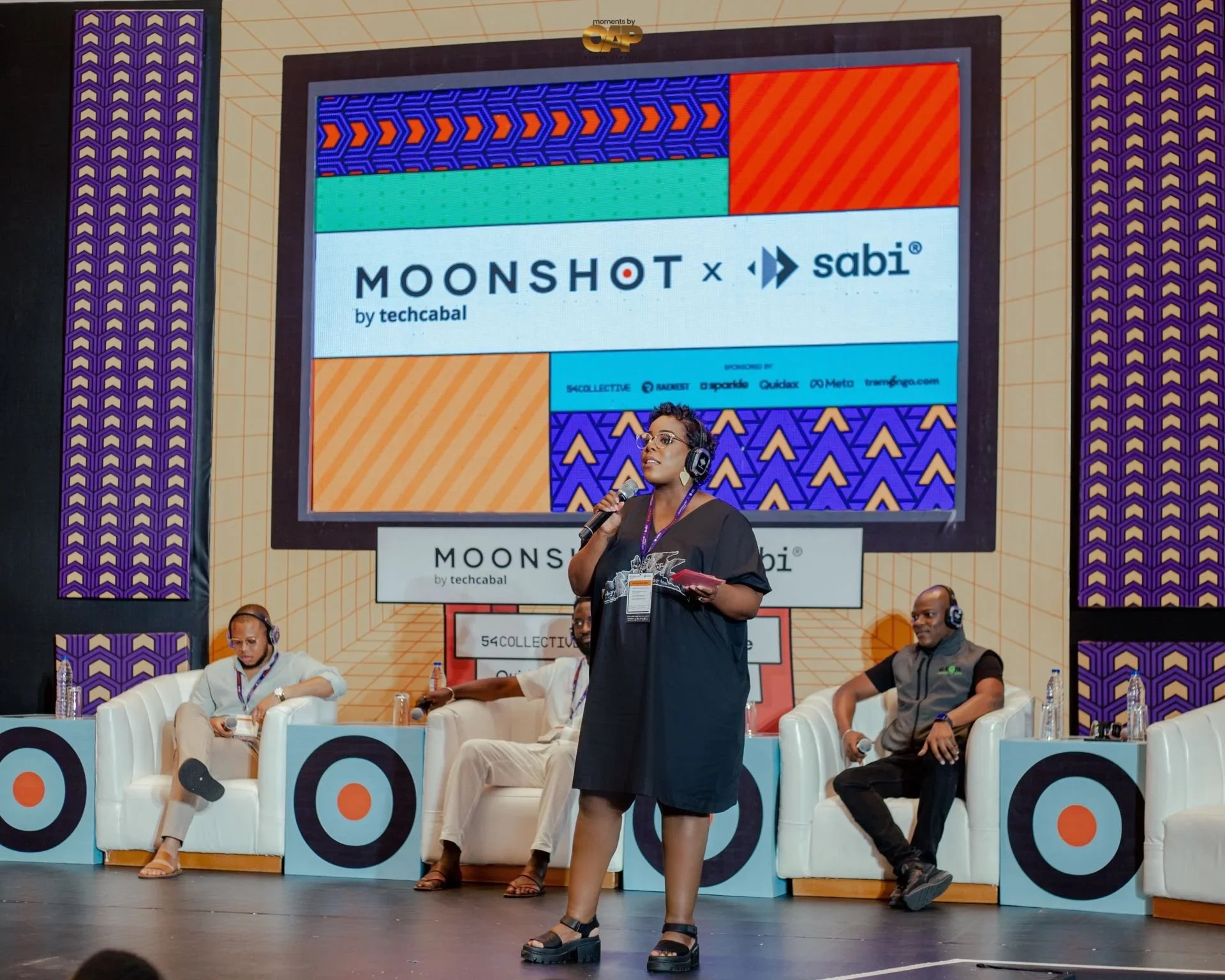 A woman standing and speaking into a microphone at a panel discussion event, with several seated speakers behind her on stage. A large screen behind them displays the event title, "MOONSHOT by techcabal," and associated logos and graphics. The stage features a vibrant, patterned background.
