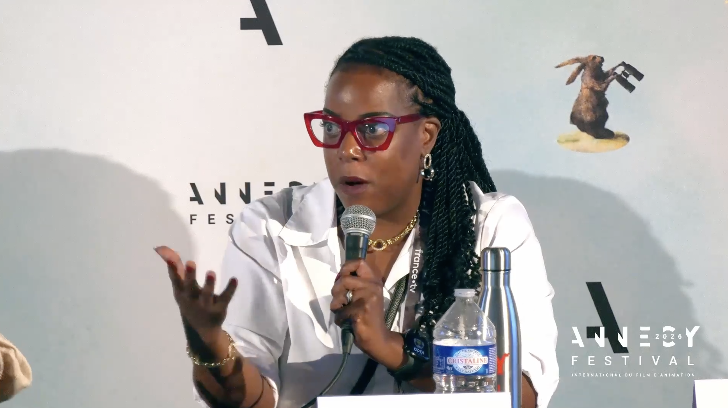 A woman with braided hair and red glasses speaking into a microphone at a panel discussion during a film festival, with a white background and water bottles in front of her.