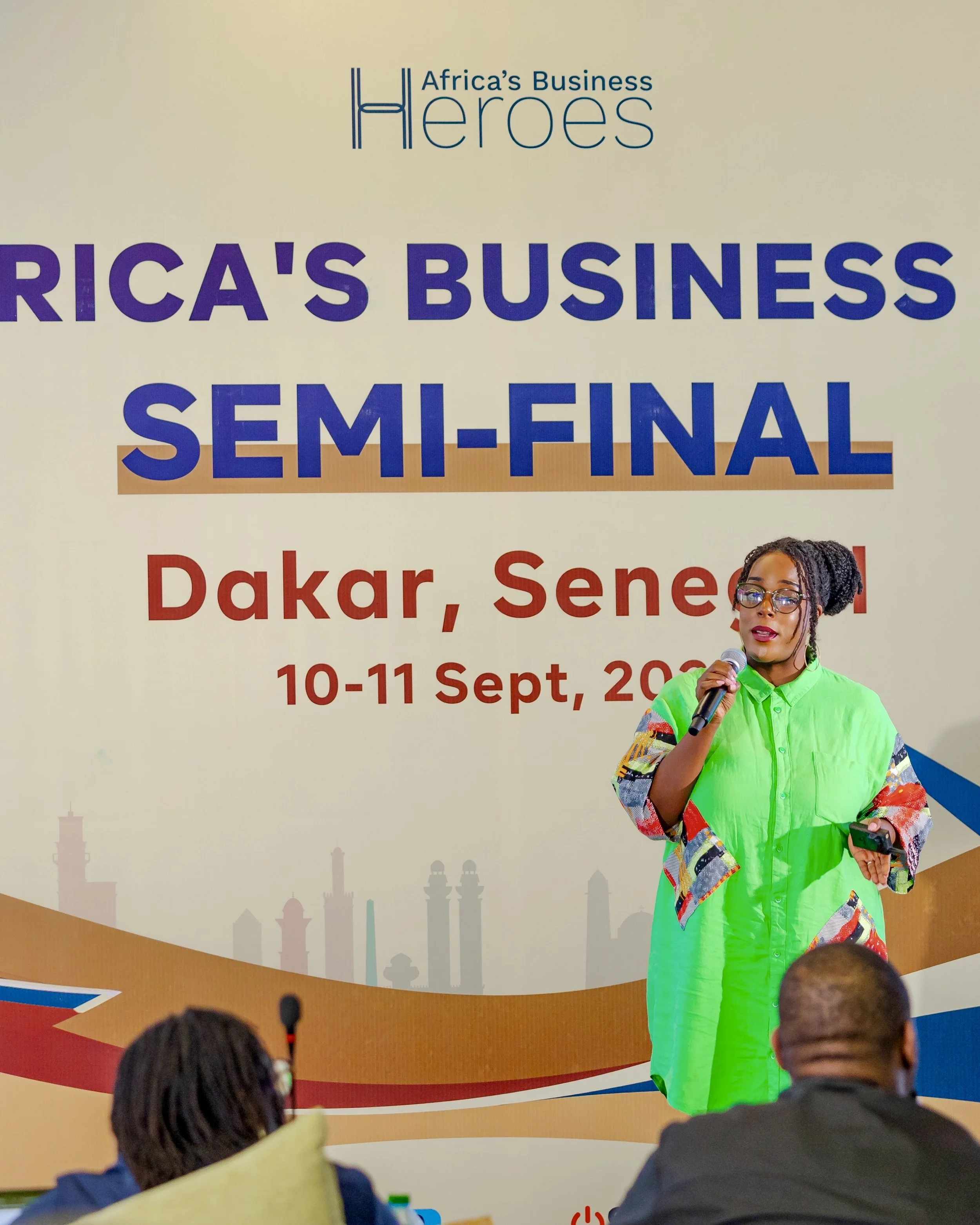 A woman speaking into a microphone on stage at a conference. She is wearing a bright green dress with colorful sleeves. Behind her is a large banner that reads "Africa's Business Heroes Semi-Final" with the location and date details.