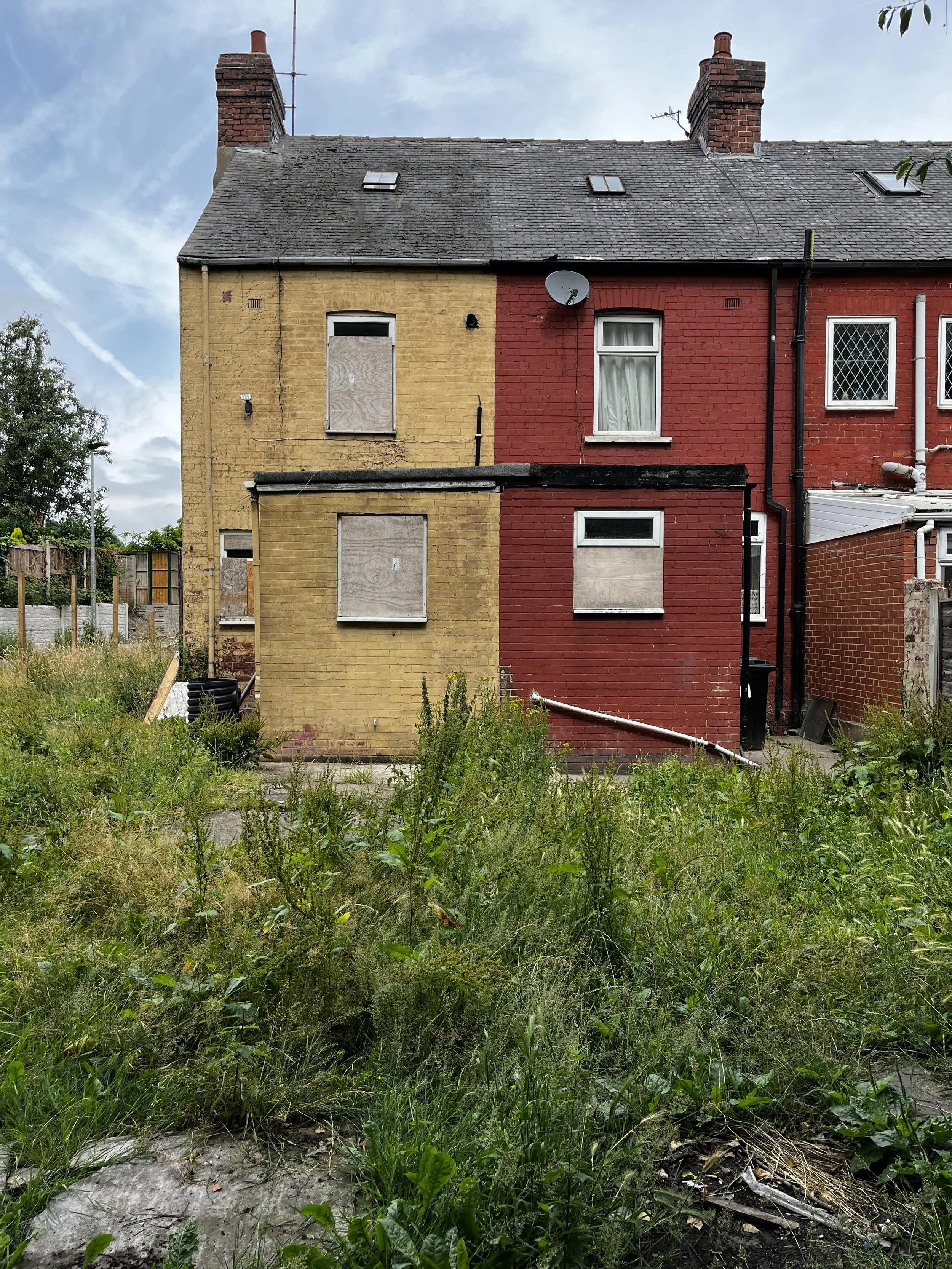 A row of houses with boarded-up windows and unkempt garden in front, overgrown with grass and weeds, under a partly cloudy sky.