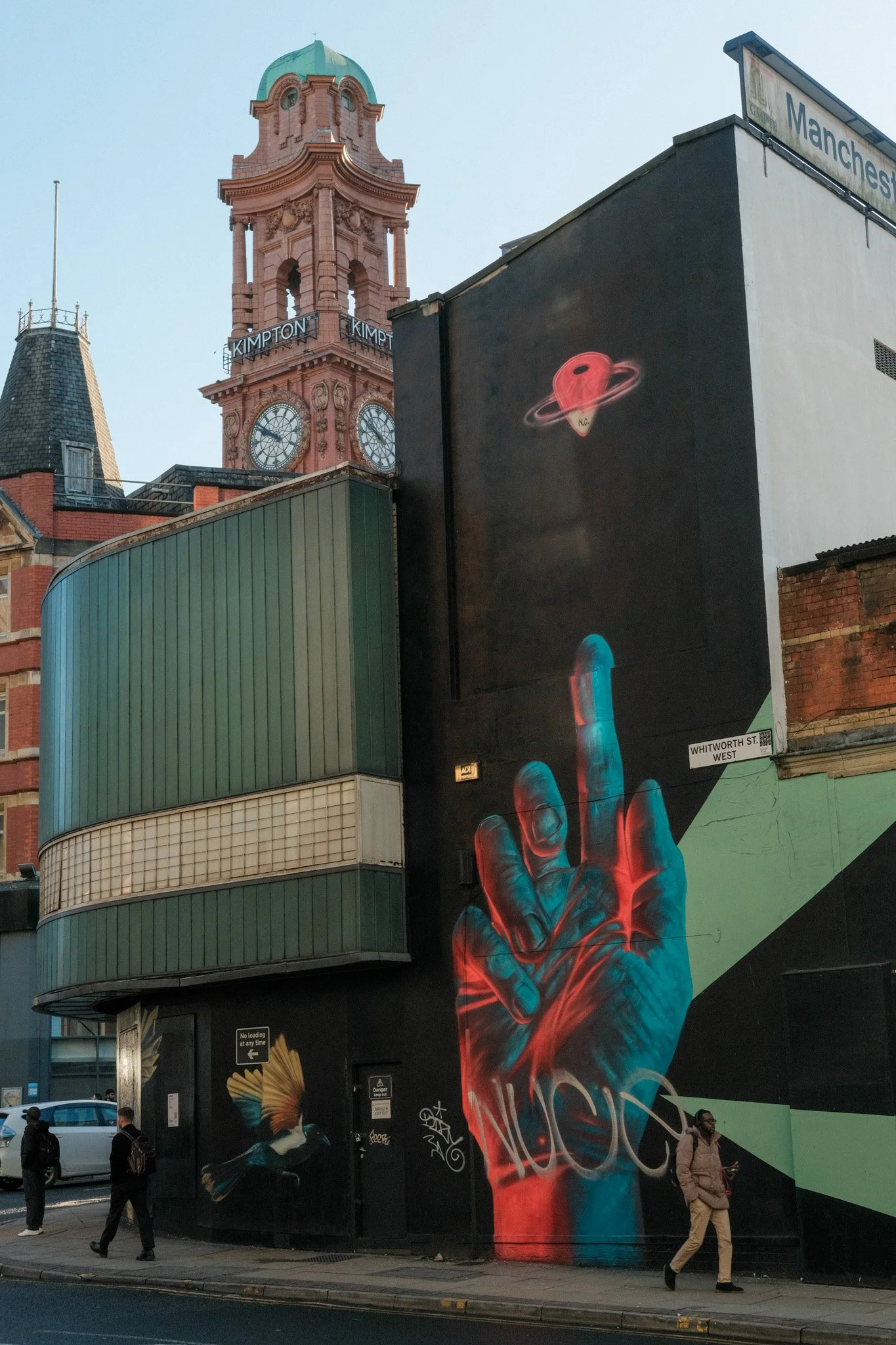 Street scene with four pedestrians, graffiti art of a red hand, a building with a clock tower, and billboard signs, including 'Kimpton' and 'Manchester'.