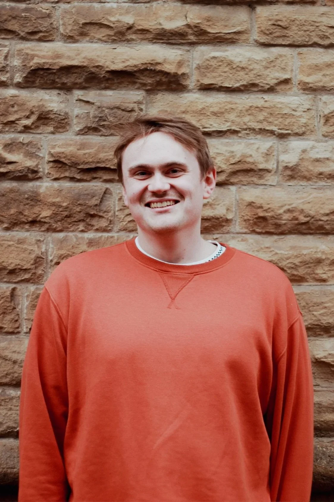 A young man with short brown hair, smiling, standing in front of a textured beige brick wall, wearing an orange sweatshirt.