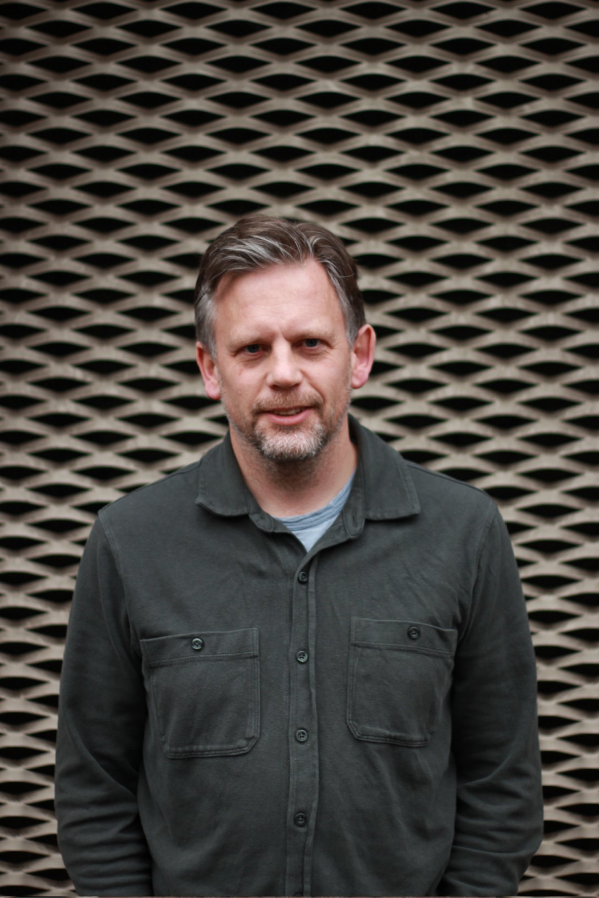 A man with short brown hair and a beard standing in front of a patterned background, wearing a dark button-up shirt.