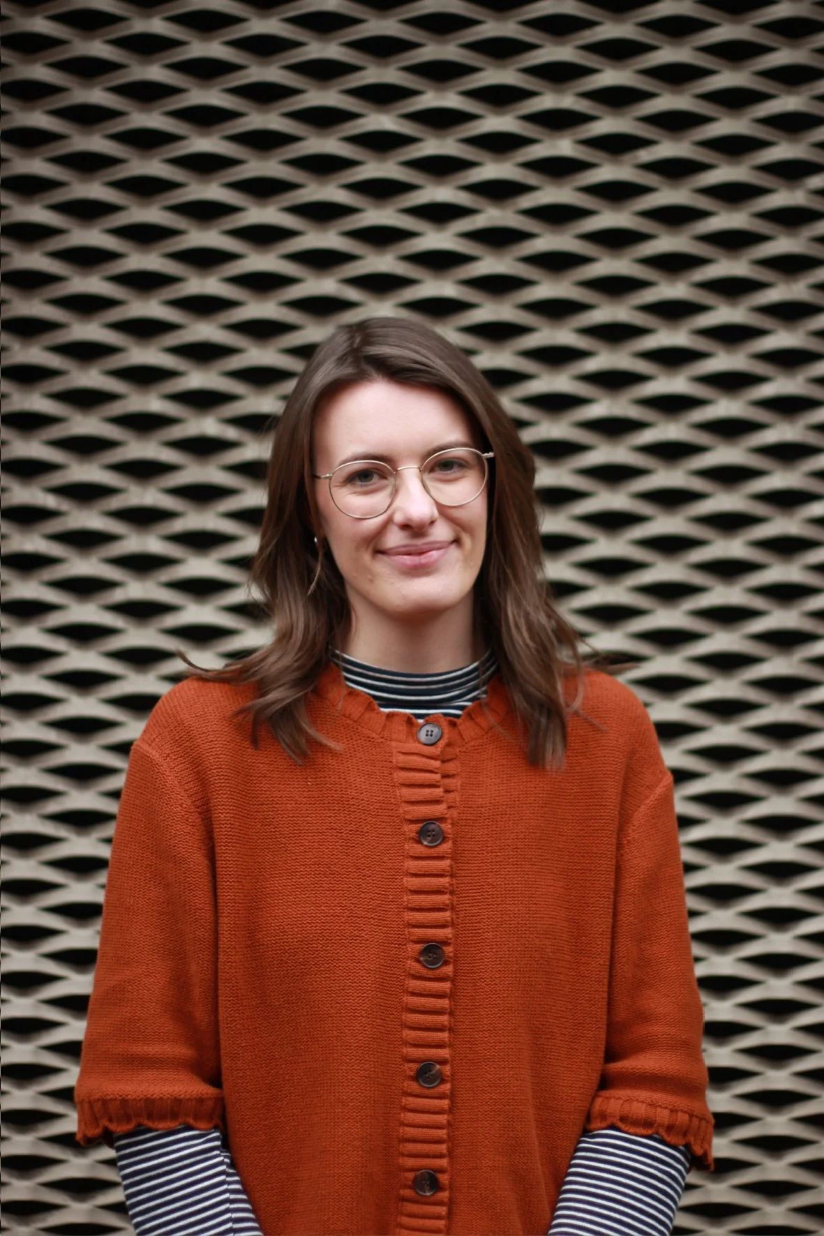 A young woman with shoulder-length brown hair and glasses, wearing an orange cardigan over a black and white striped shirt, standing in front of a black lattice background.