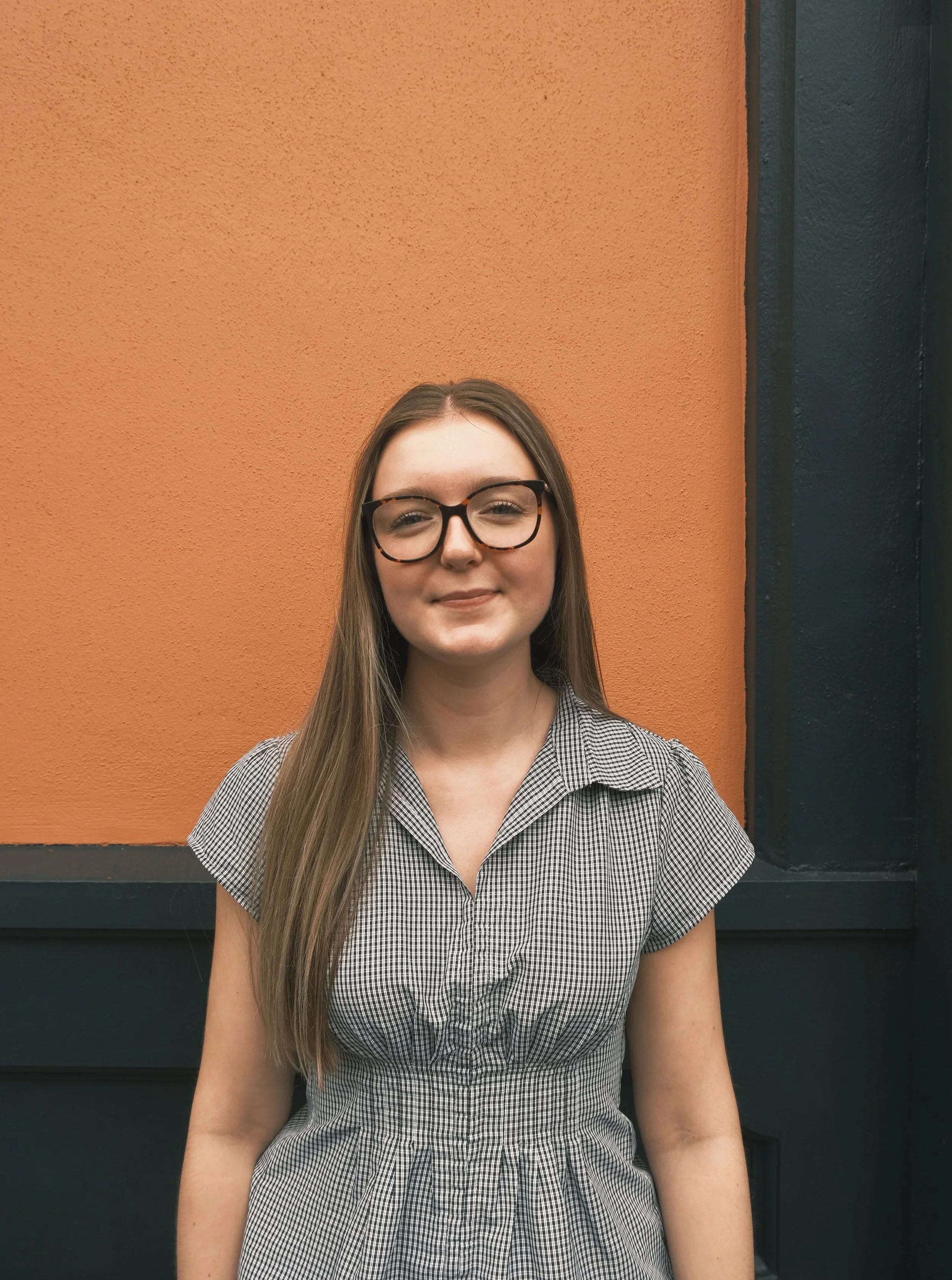 A young woman with long brown hair, wearing glasses and a checkered dress, sitting against an orange and black wall.