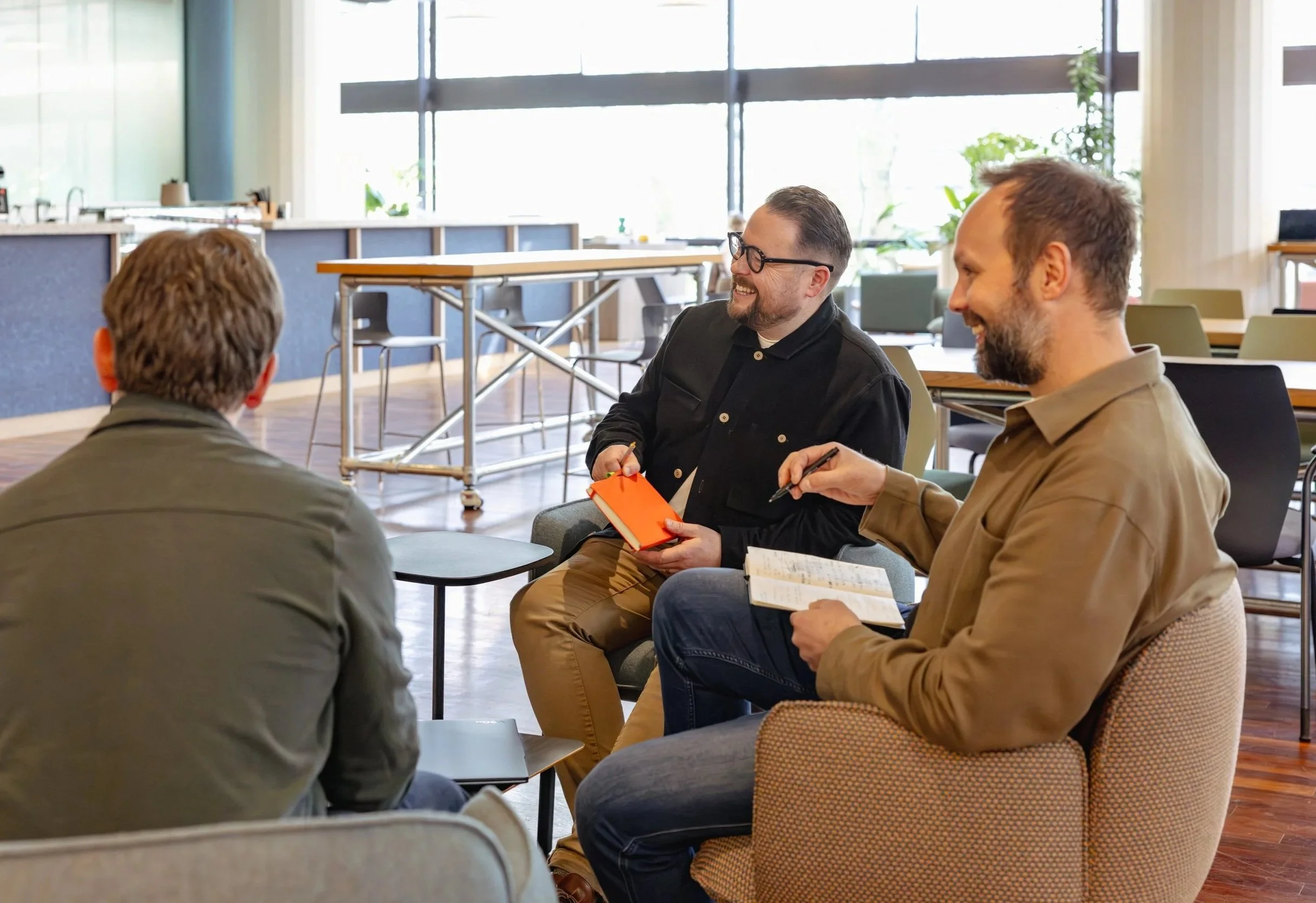 Four men sitting in a circle having a conversation in a bright, modern room with large windows and plants.