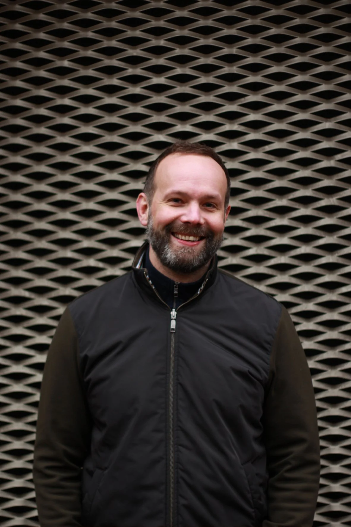 Smiling man with a beard and short hair standing in front of a patterned wall.